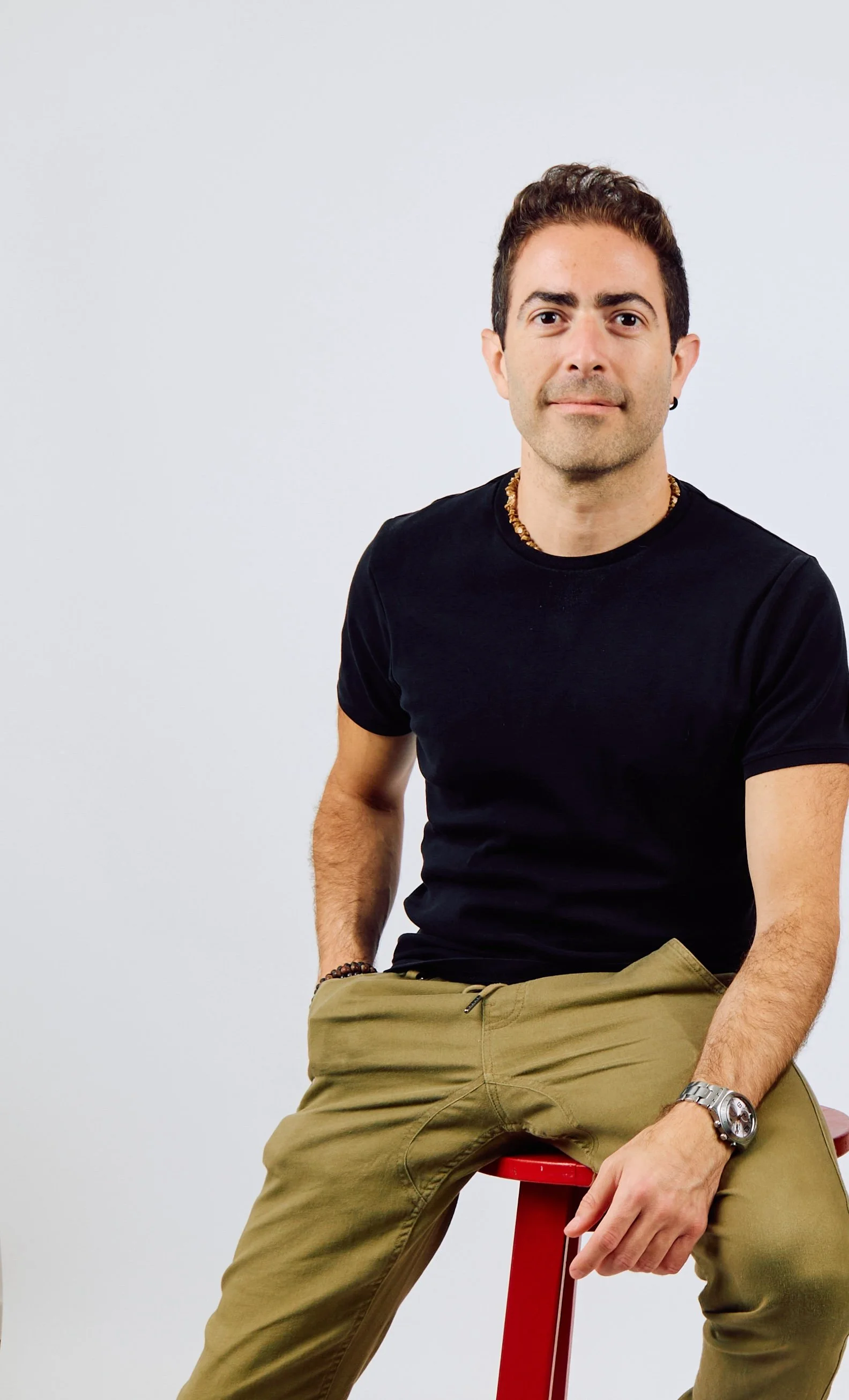 A man with short brown hair, fair skin, wearing a black T-shirt, khaki pants, and accessories including a watch, wristband, and beaded necklace, sitting on a red stool against a plain white background.