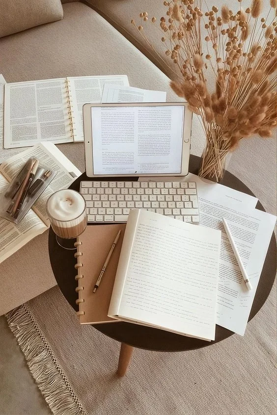A round black table with an open notebook, pen, and a tablet, surrounded by open books, a candle, a pen container, and a vase of dried flowers, all on a beige sofa and carpet.