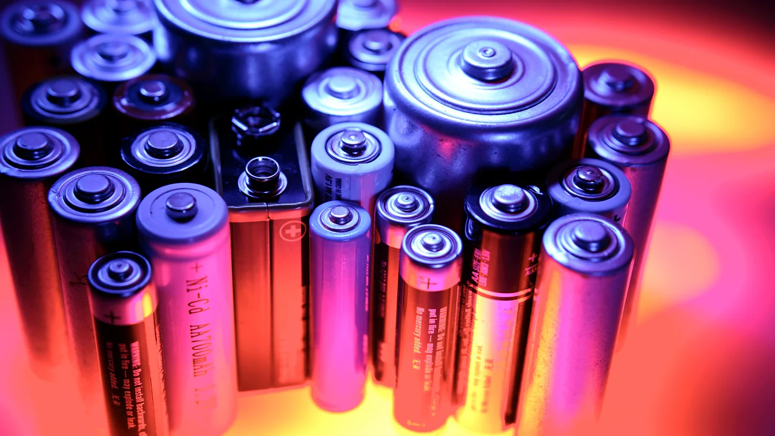 Close-up of various batteries, including AA, AAA, and other cylindrical batteries, arranged in a cluster with colorful lighting.