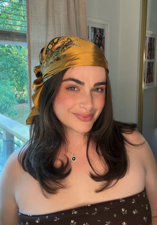 A woman with long dark hair wearing a gold patterned headscarf and a strapless floral dress, smiling in front of a window with trees outside.