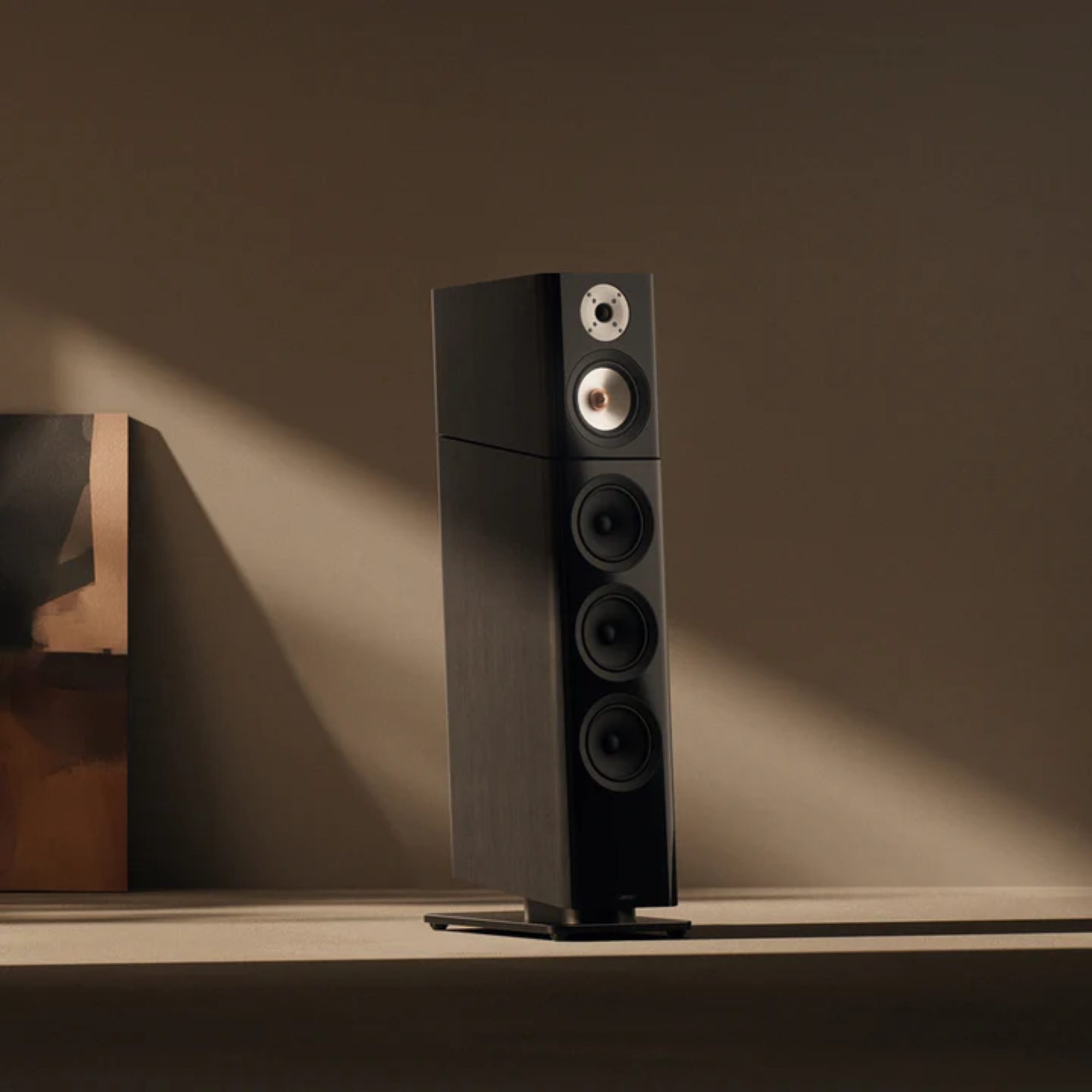 A tall black speaker with silver accents sits on a white shelf against a beige wall from Jamo