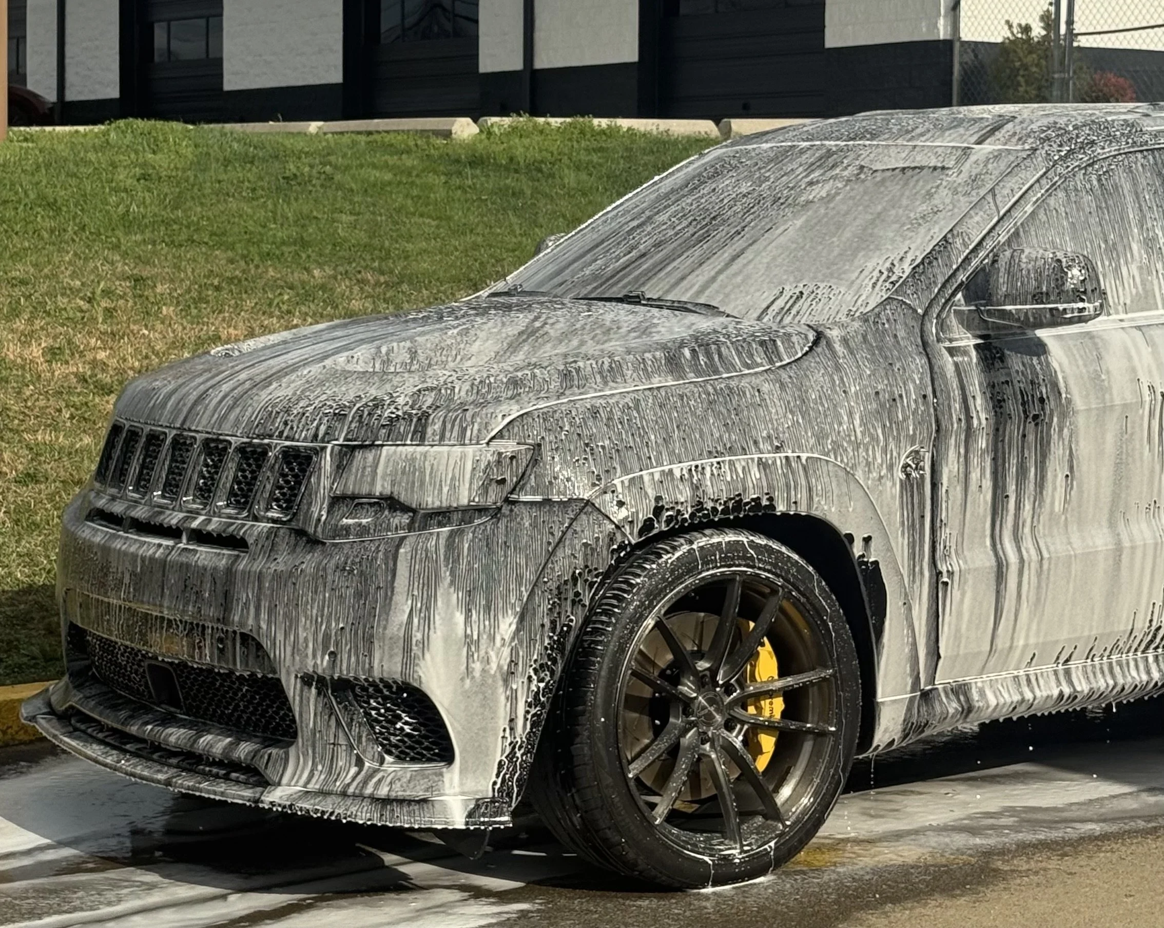 A black luxury car covered in soap suds during a car wash outside on a parking lot, with a grassy area and a building in the background.