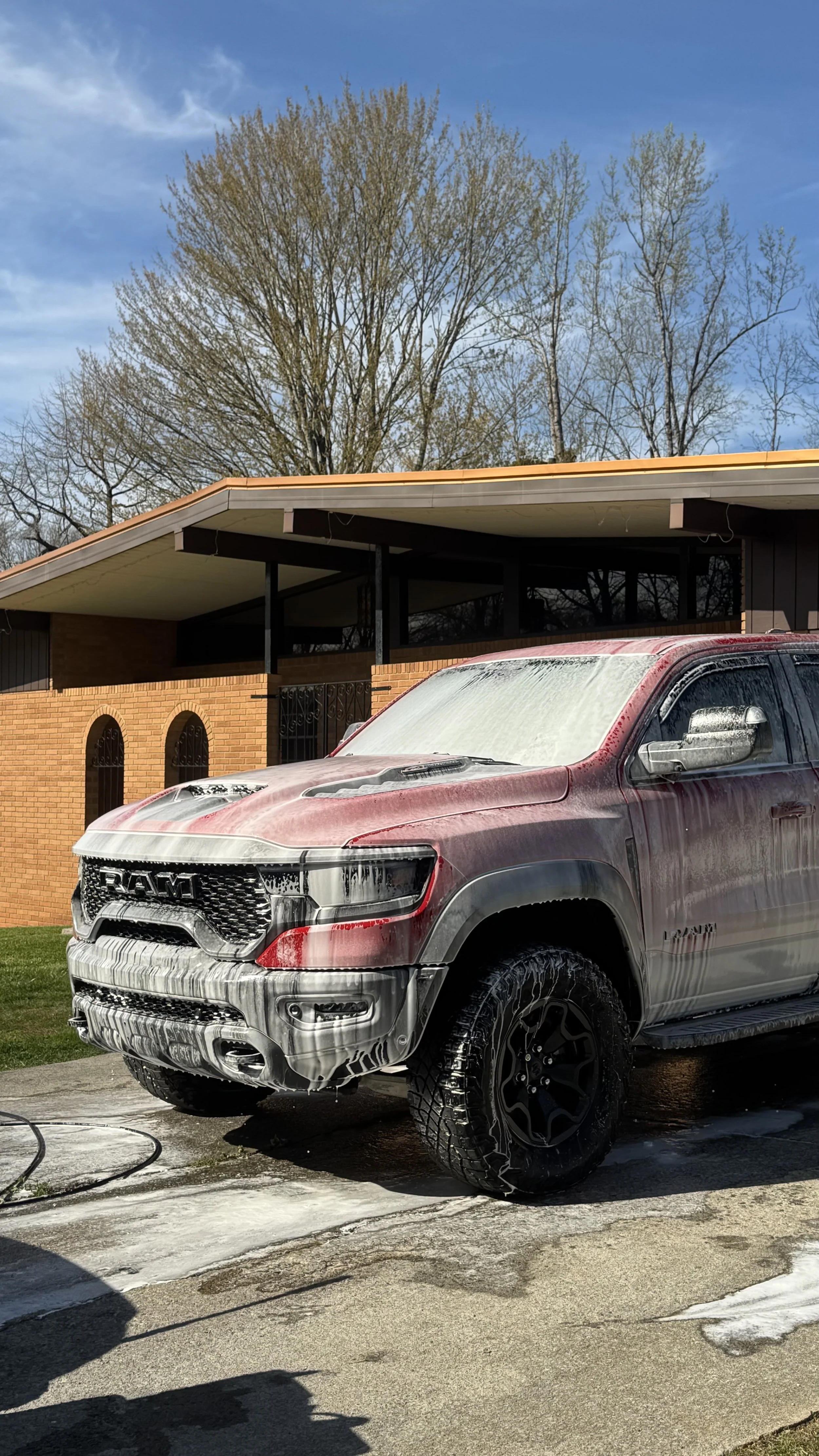A red RAM truck covered in soap suds during a washing process, parked on a driveway in front of a brick house with a sloped roof, with trees and a blue sky in the background.