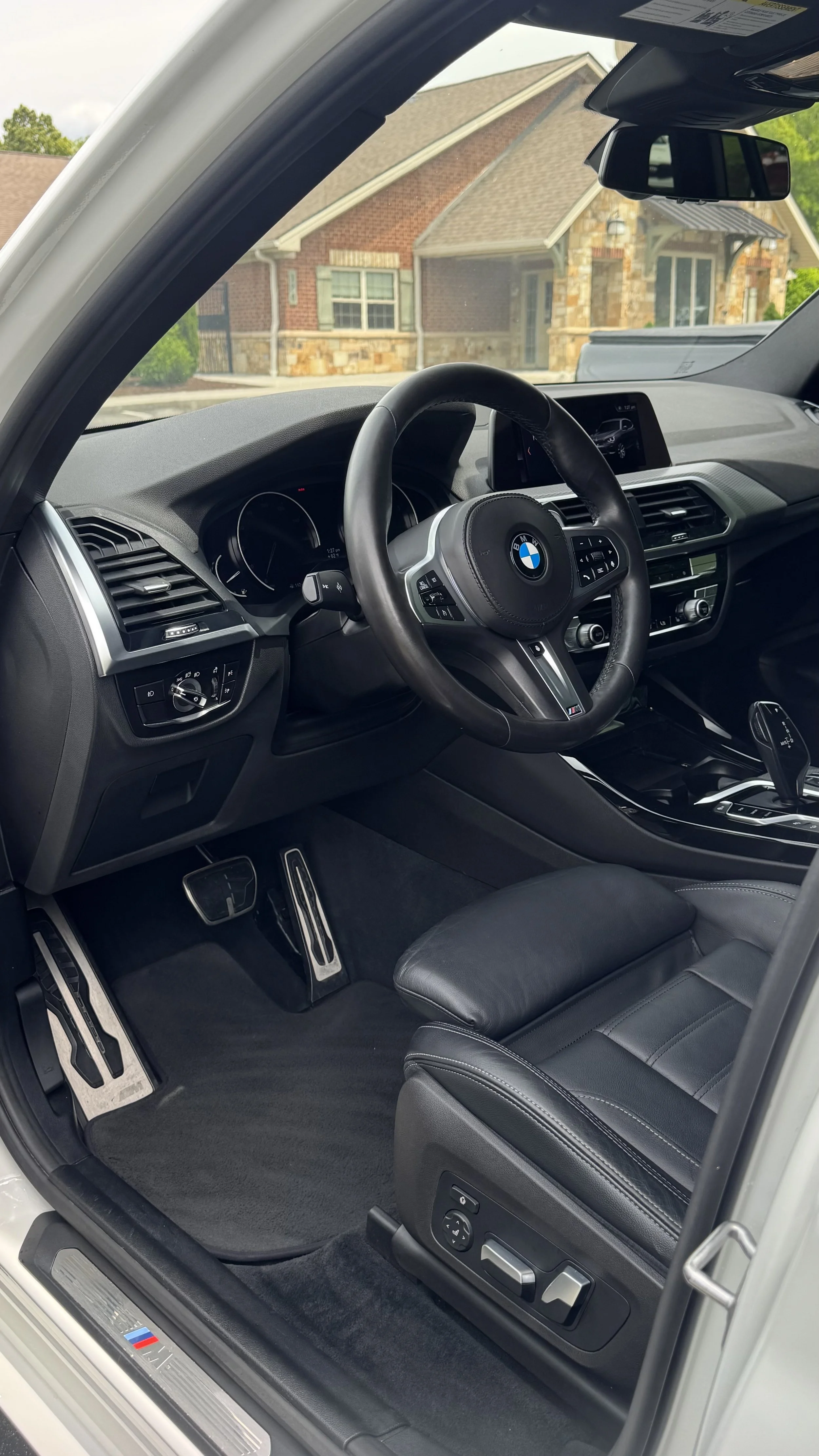 Interior of a BMW vehicle showing the steering wheel, dashboard, gear shifter, black leather seat, and foot pedals, with view of a house outside through the windshield.