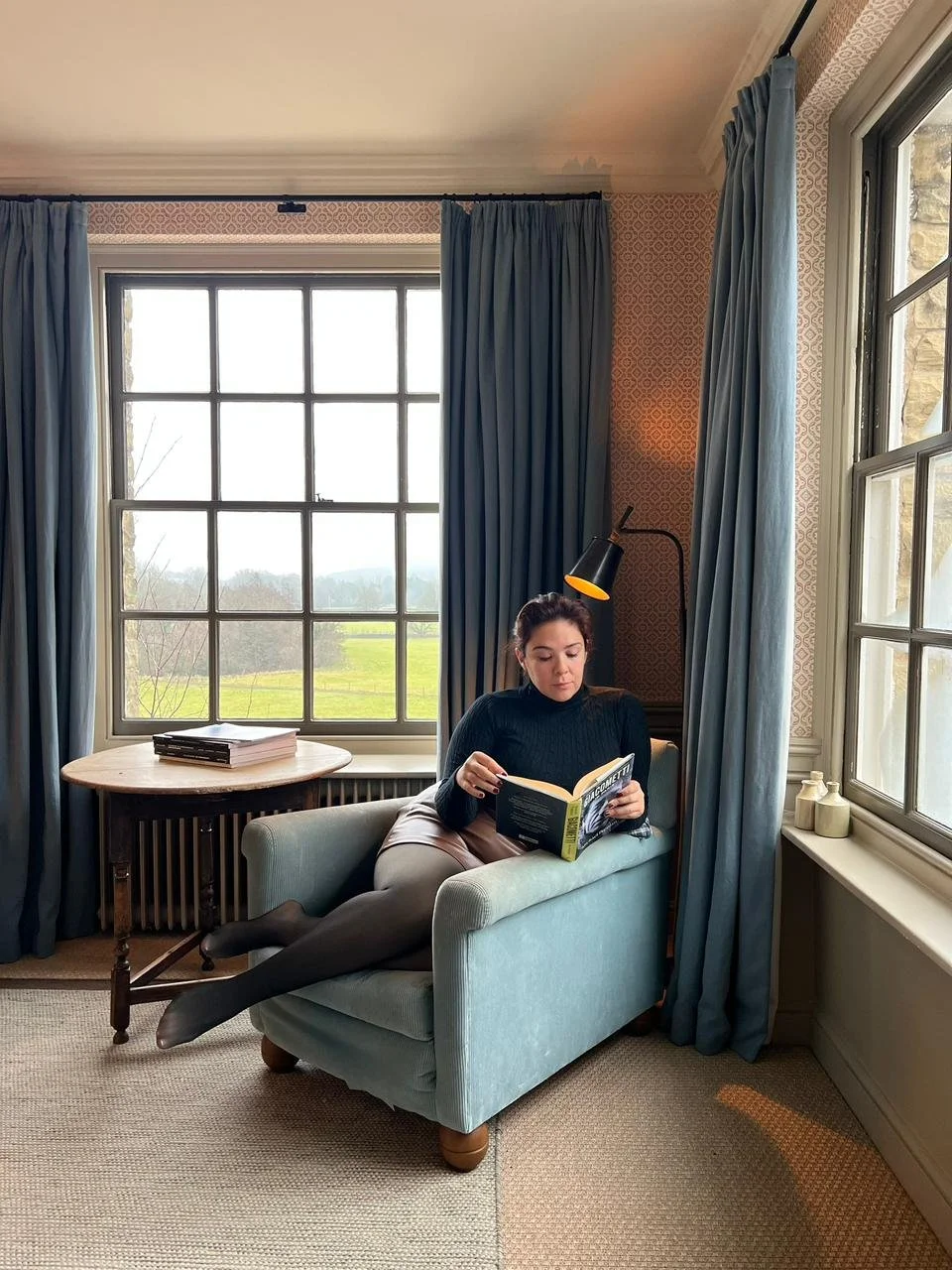 A woman in a black sweater and gray skirt reading a book while lying on a light blue armchair near large windows with blue curtains, in a room with patterned wallpaper and a side table with books, with countryside view outside. Content creator.