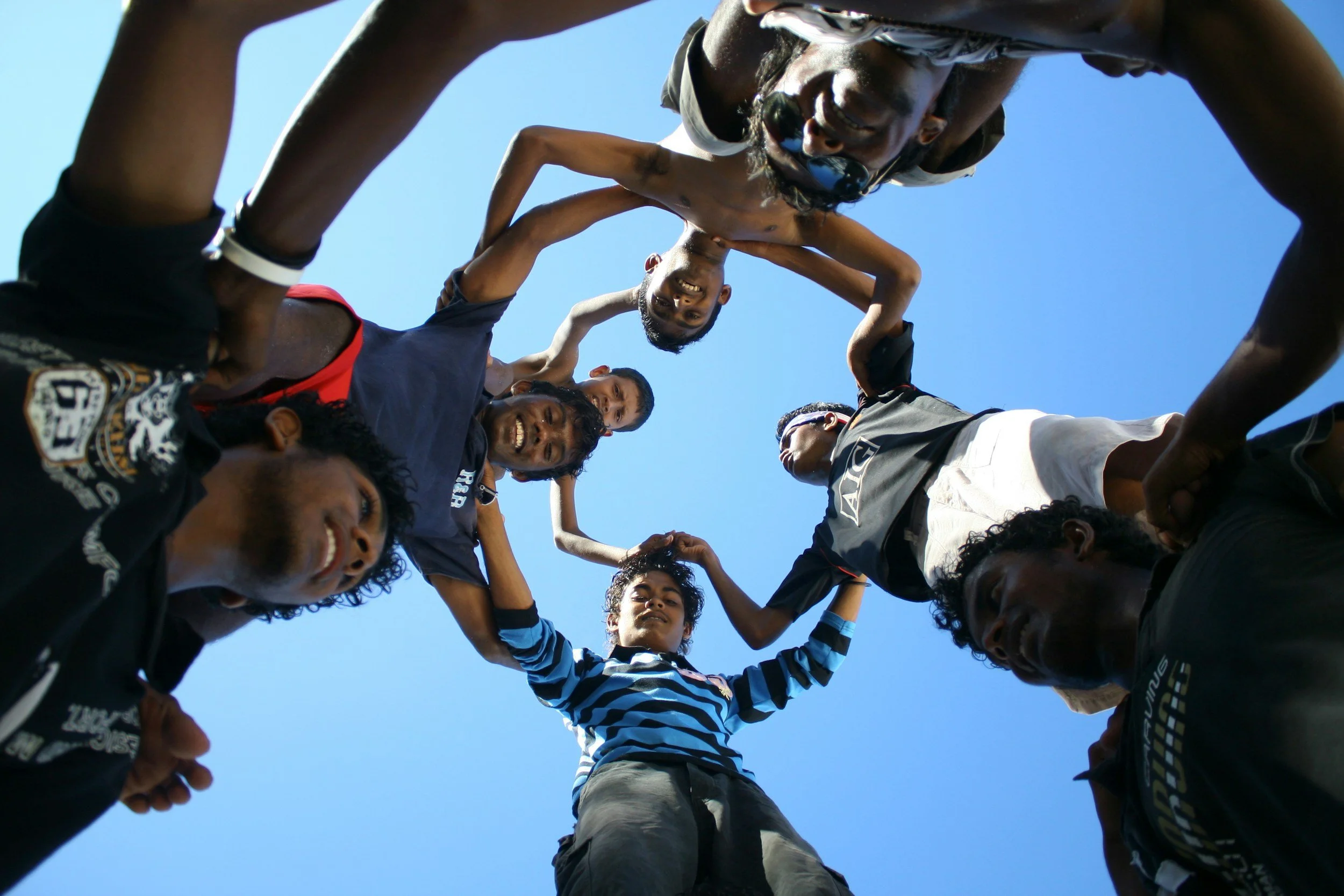 A group of young people forming a human tower, looking down at the camera against a clear blue sky.