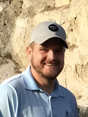A smiling man wearing a light-colored baseball cap and a blue collared shirt, outdoors in front of a rocky background.