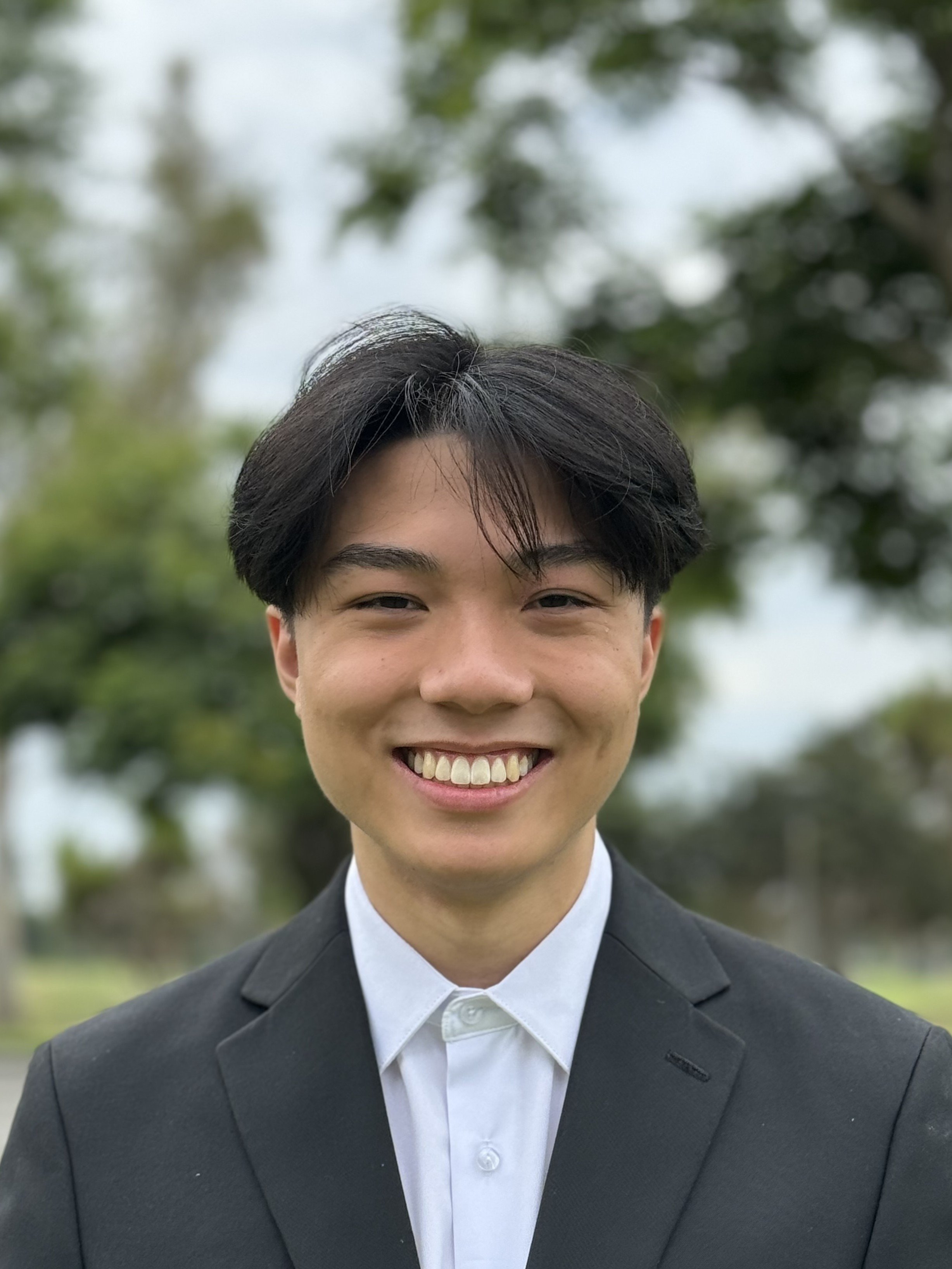Close-up of a young man smiling, wearing a black suit and white shirt, outdoors with blurred trees in the background.