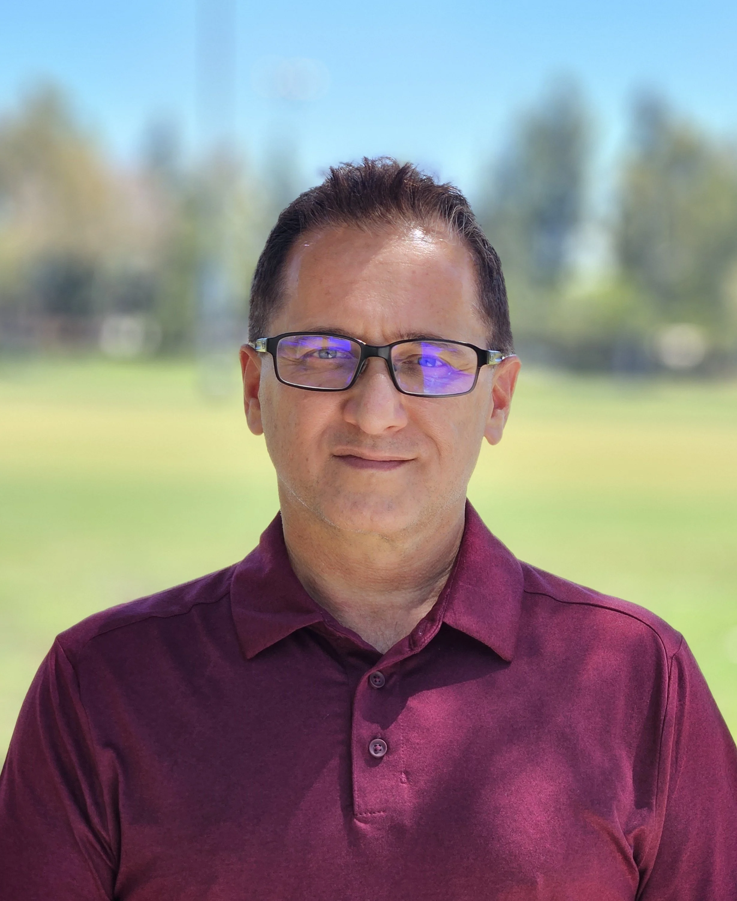 Photograph of a man wearing glasses and a maroon dress shirt, outdoors with blurred trees and sky in the background.