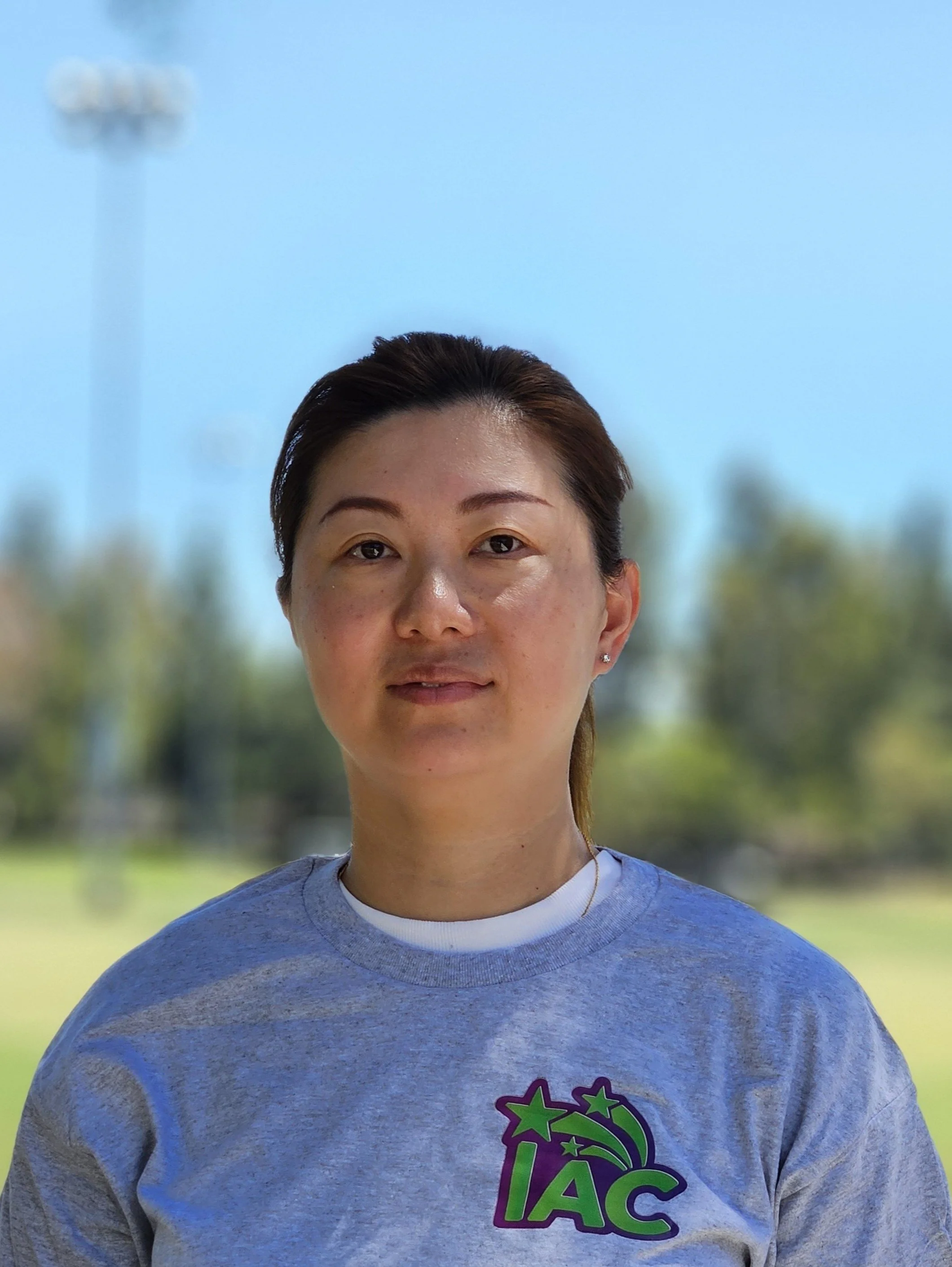 Close-up of a young woman with dark hair, wearing a gray T-shirt with a colorful logo, outdoors on a sunny day with blurred trees and blue sky in the background.