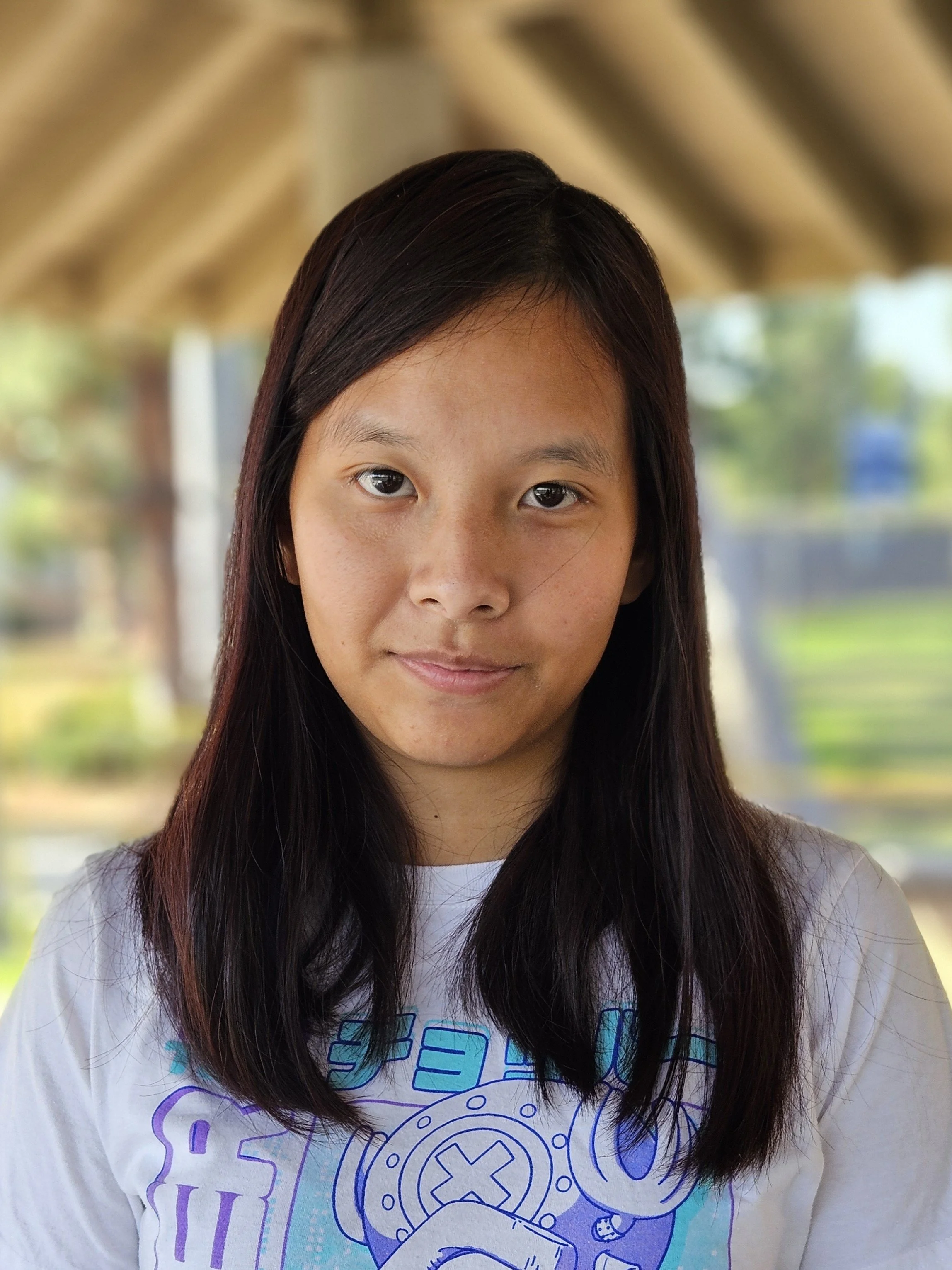 A young woman with long dark hair, wearing a white t-shirt with blue graphics, outdoor setting with blurred trees and wooden structure in the background.