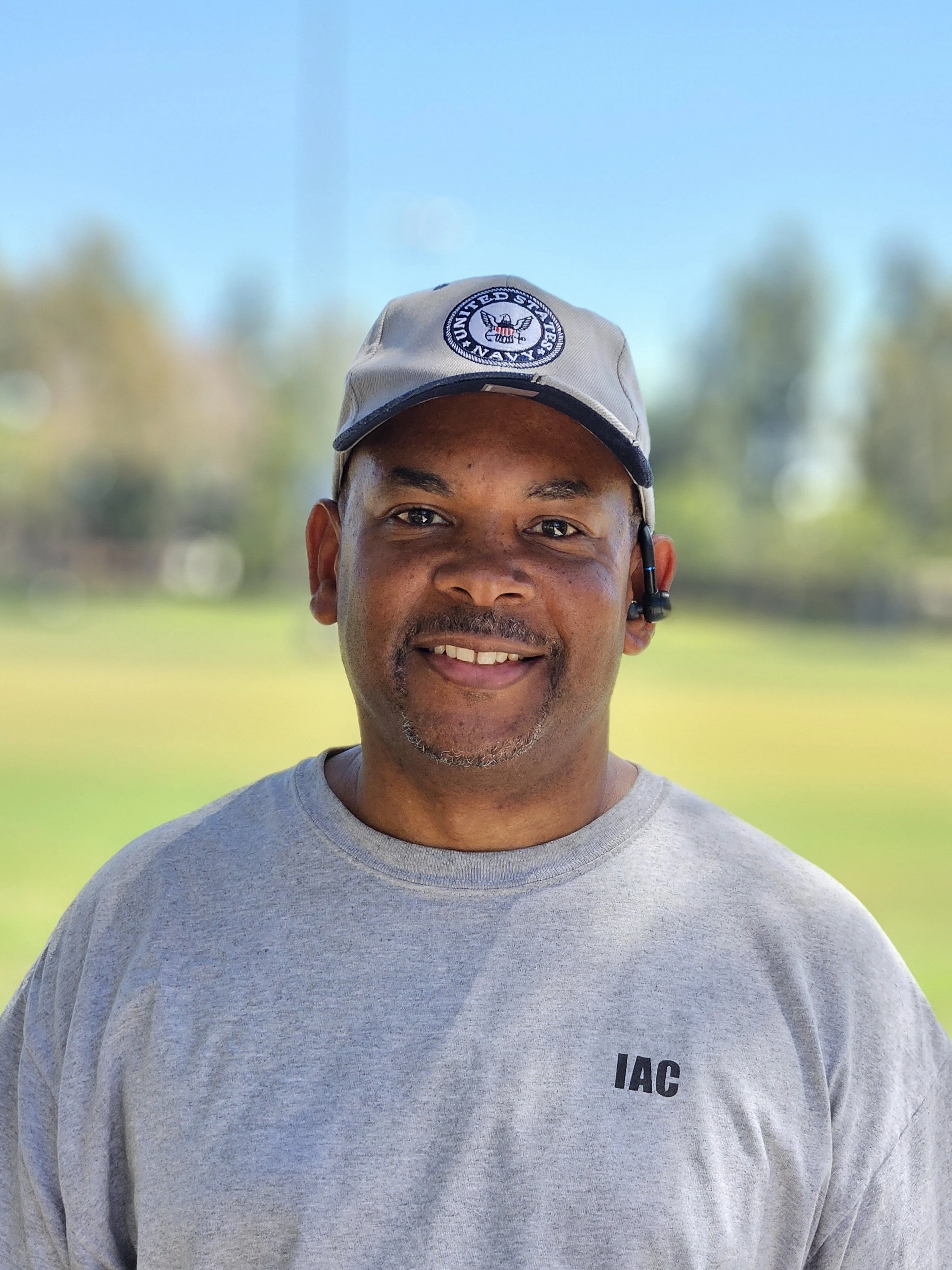 Close-up of a smiling man wearing a gray baseball cap with the United States Navy Seal and a gray sweatshirt with 'IAC' printed on it, standing outdoors with a blurred background of trees and sunlight.
