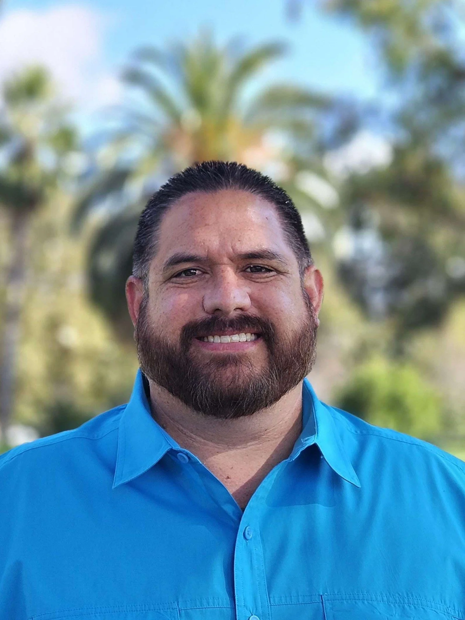 A smiling man with a trimmed beard and mustache, wearing a blue collared shirt, standing outdoors with blurred palm trees and greenery in the background.