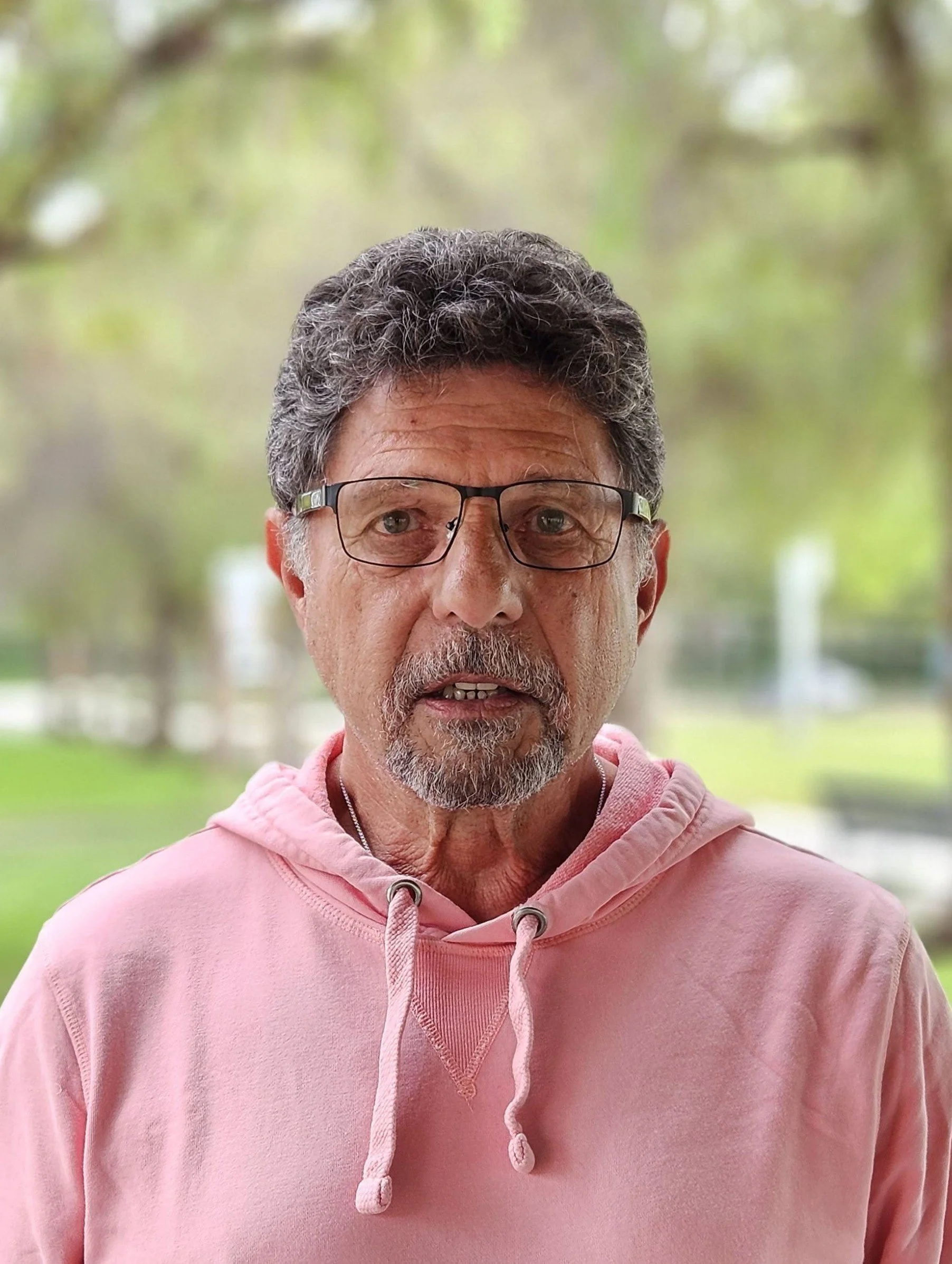 Close-up of a middle-aged man with glasses, gray curly hair, and a mustache, wearing a pink hoodie outdoors with a blurred green background.