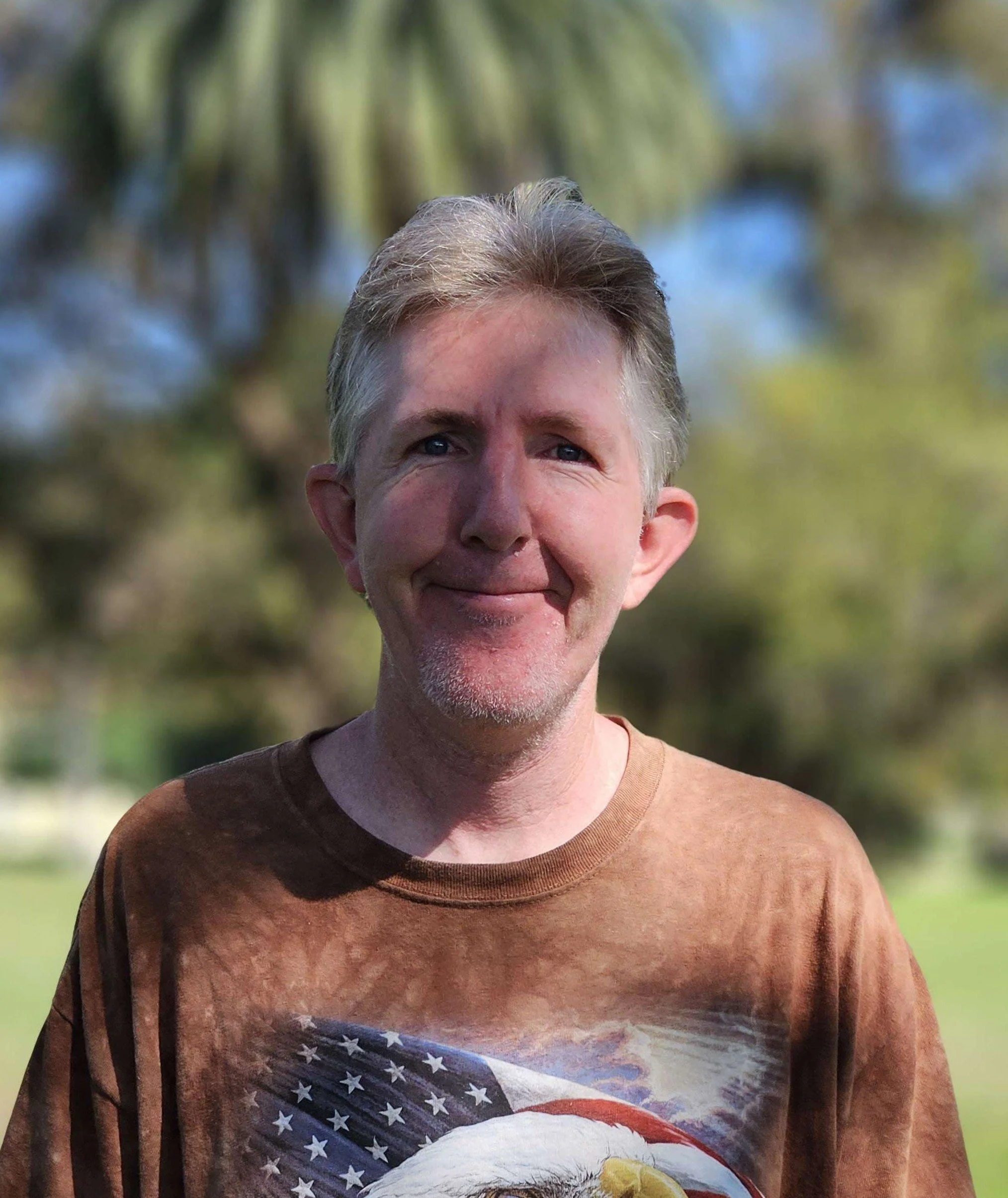 Close-up of a middle-aged man with gray hair, wearing a brown t-shirt with an American flag eagle graphic, outdoors with blurred trees and sky in background.