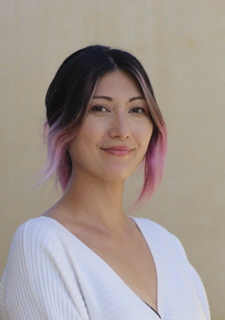A woman with short black hair with pink tips, smiling, wearing a white v-neck top, standing against a plain beige wall.