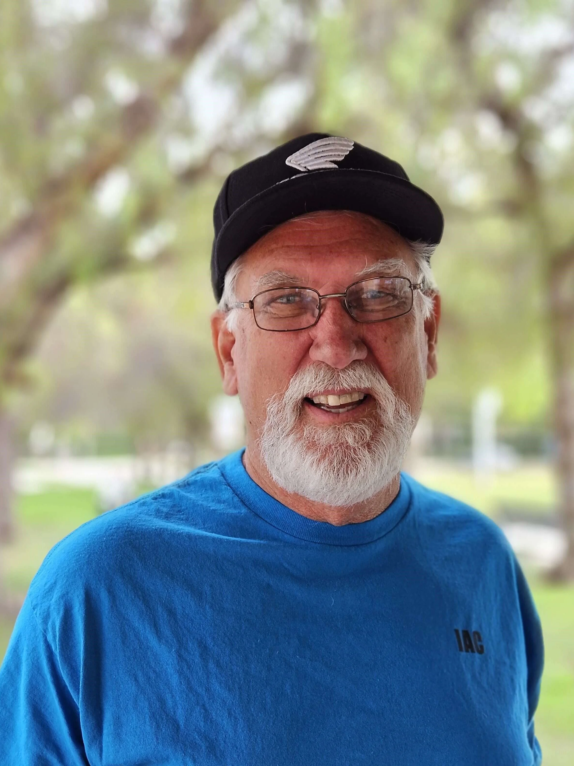 A close-up of an elderly man with white hair, a white beard, glasses, wearing a black cap with a white logo, and a bright blue shirt with the partial text 'IAG' visible. The background is a blurry outdoor setting with trees.