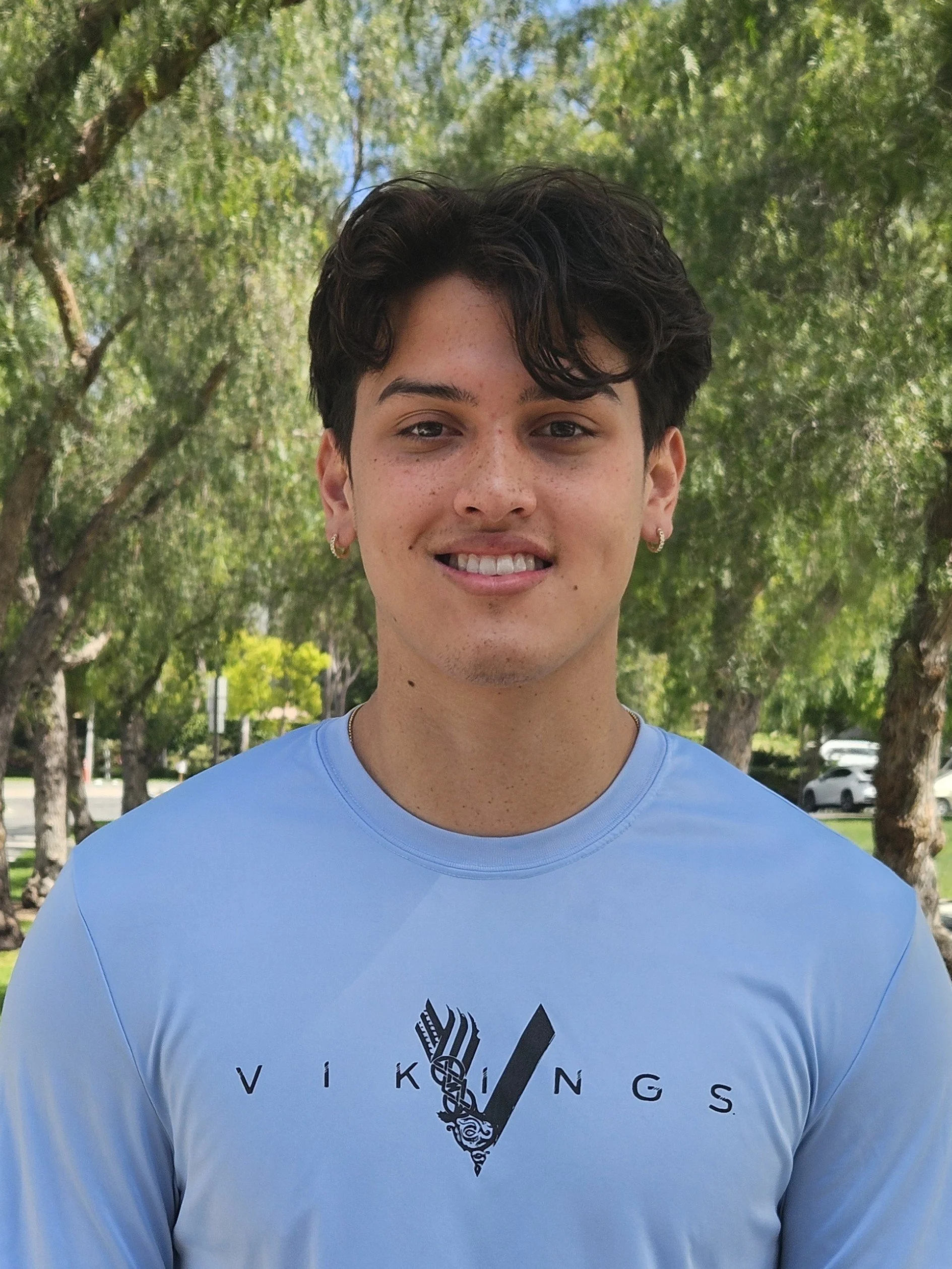 Close-up of a young person with short dark hair wearing a light blue T-shirt with a Viking helmet logo and the words Viking Vessels, standing outdoors with trees and cars in the background.