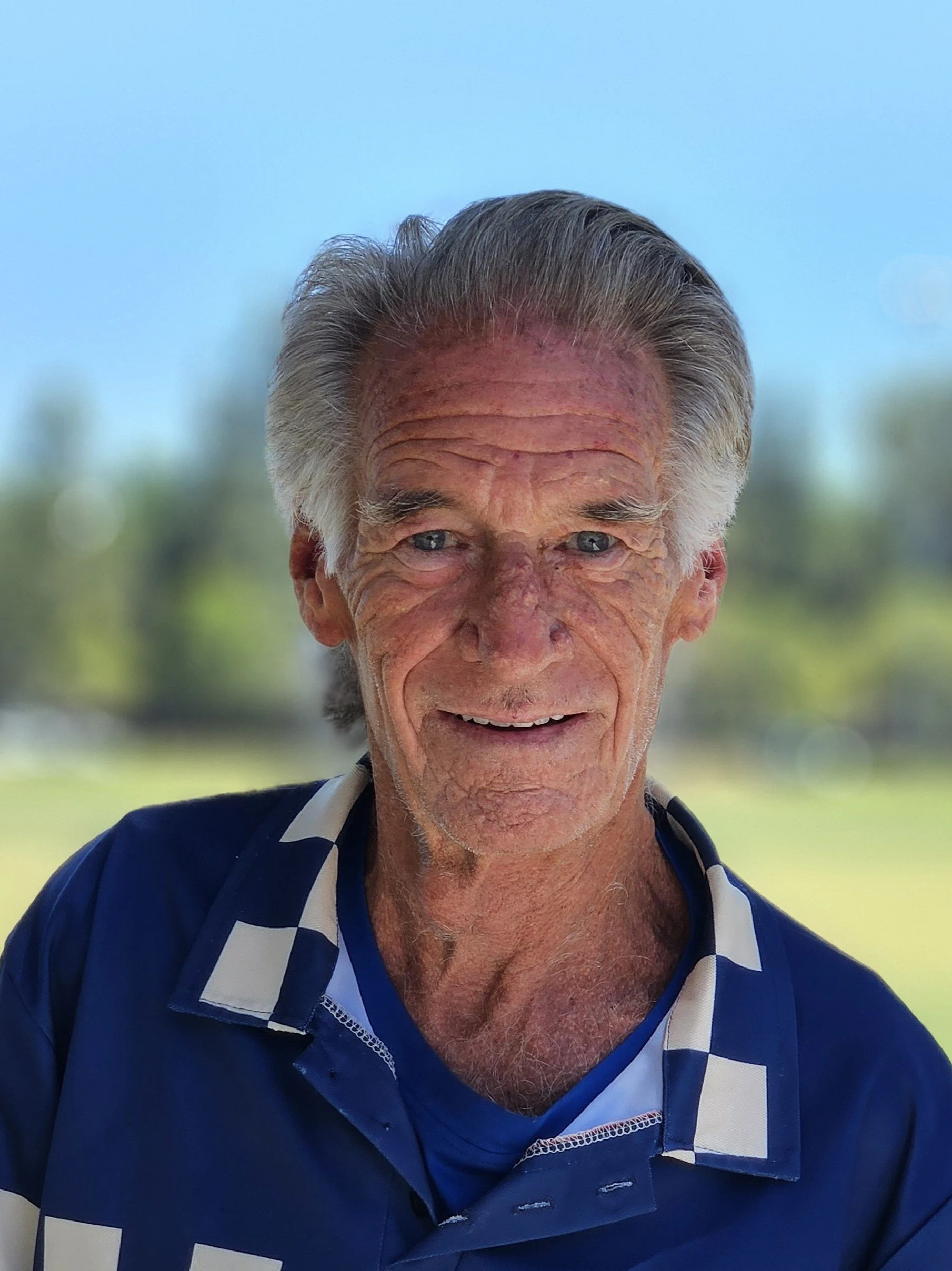 Close-up of an elderly man smiling outdoors, wearing a blue and white checkered sports shirt, with short gray hair and a background of blurred green trees and blue sky.