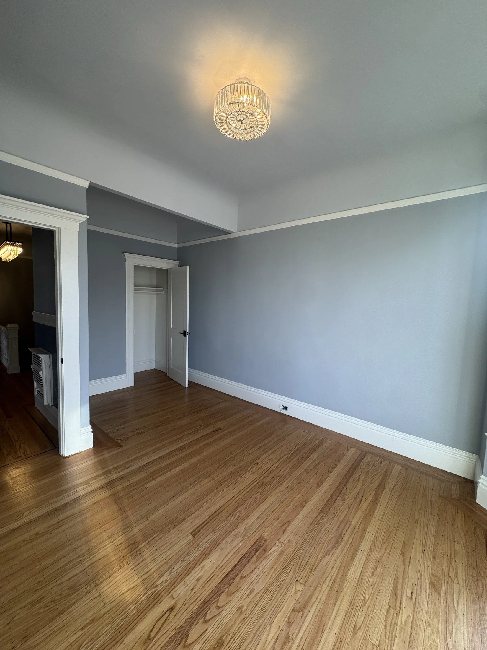 An empty room featuring light blue painted walls, white trim, and hardwood floors. There is an open closet door and a small hallway visible.