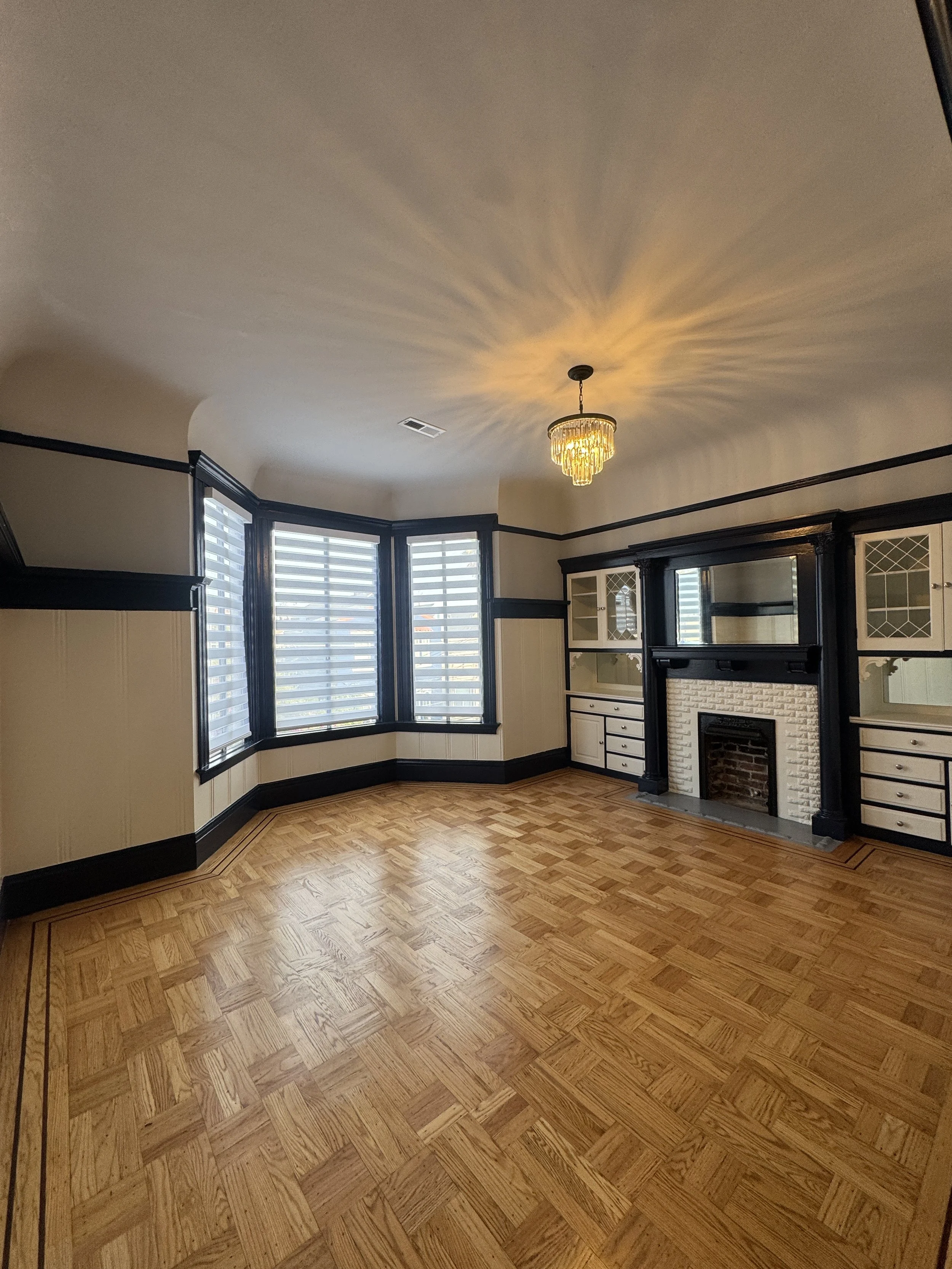Living room with hardwood floors, a fireplace surrounded by black cabinetry, and a ceiling with a decorative chandelier. Large windows with blinds let in natural light.
