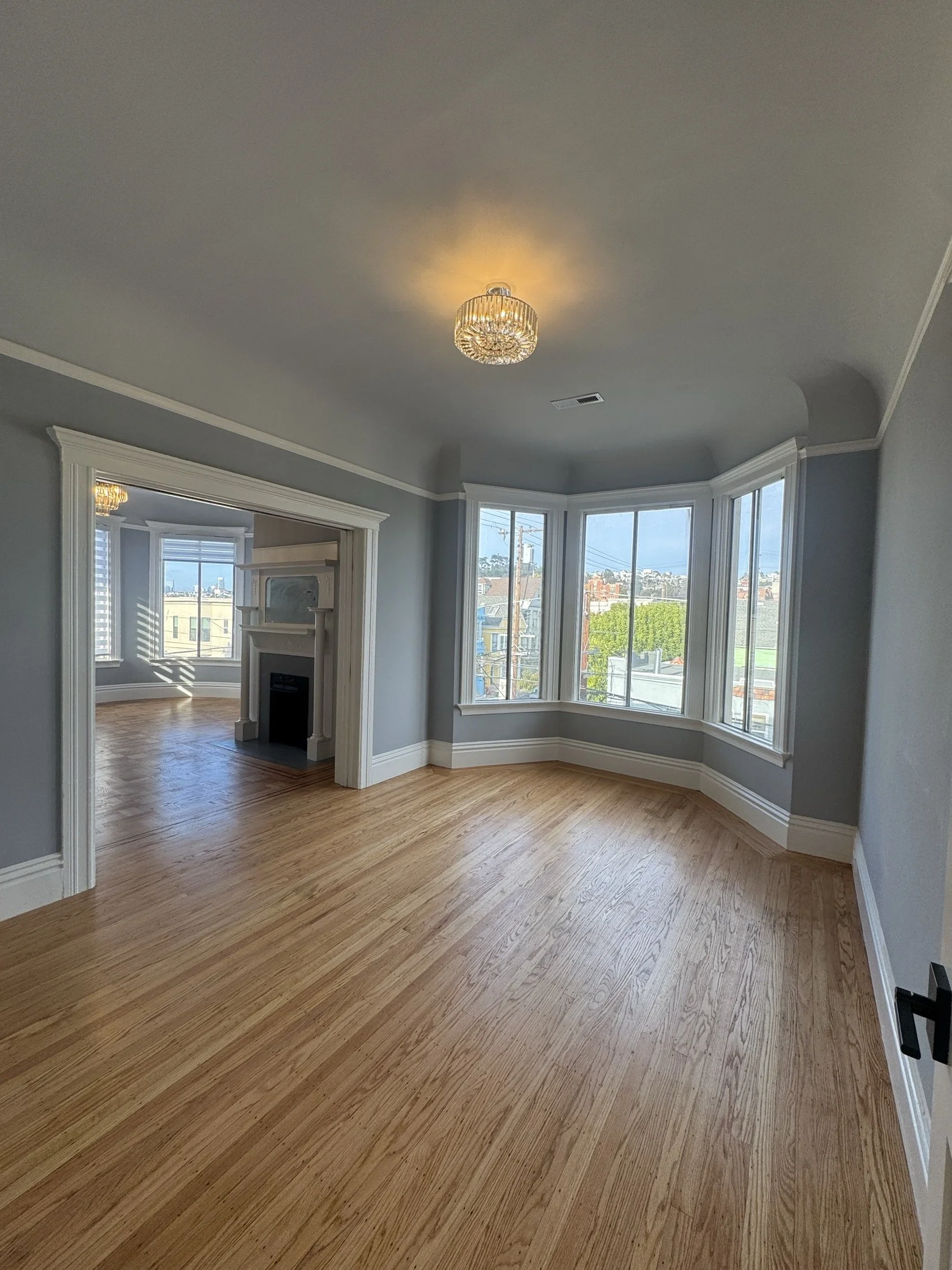 Empty living room with hardwood floors, large bay windows, a ceiling light fixture, and a fireplace in the adjacent room.