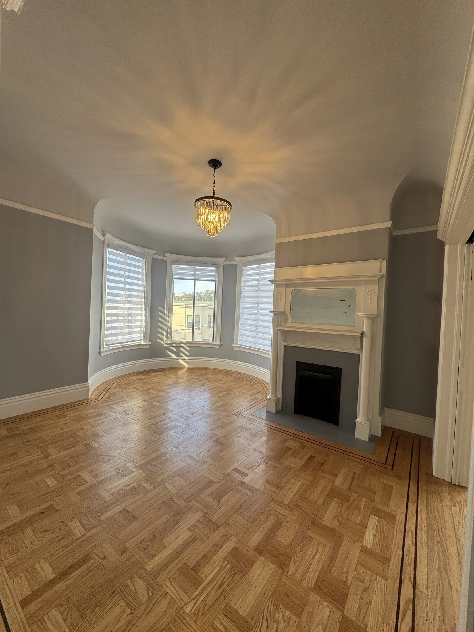 Living room with large bay window, hardwood parquet flooring, gray walls, fireplace with white mantel, and a decorative chandelier lighting fixture.