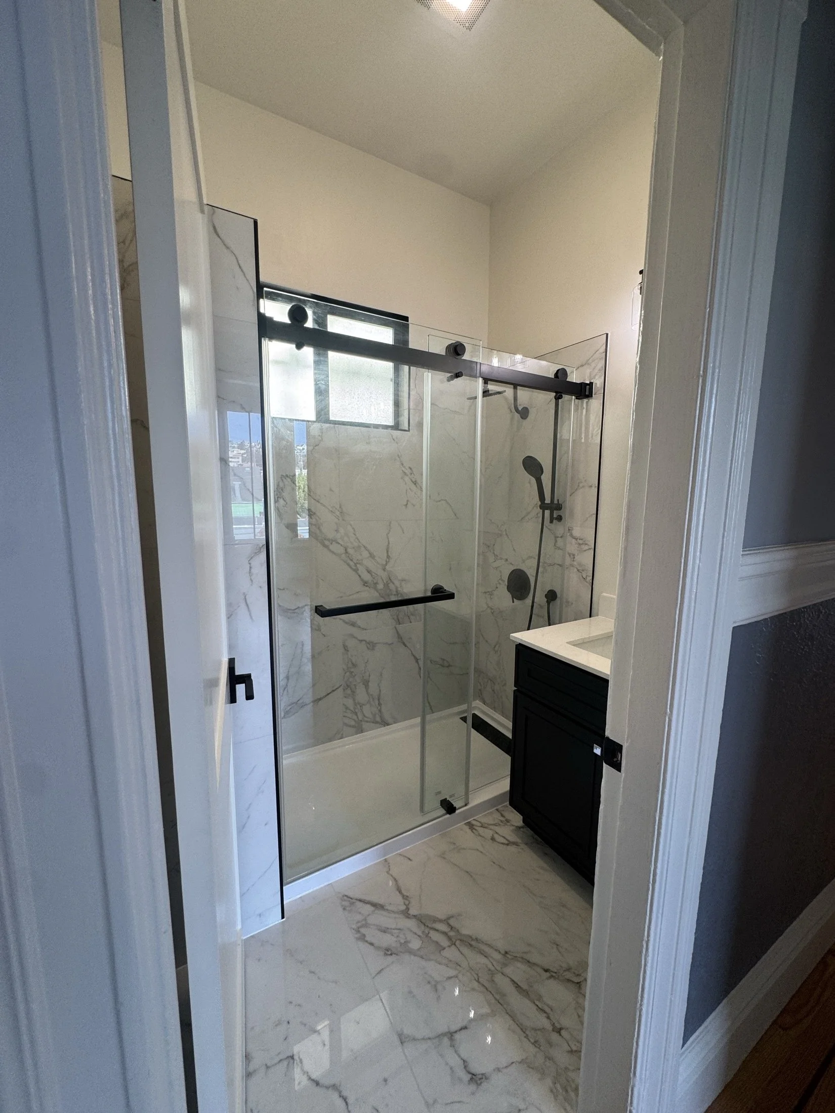 Modern bathroom with marble flooring and walls, glass shower enclosure, black vanity, and a window.