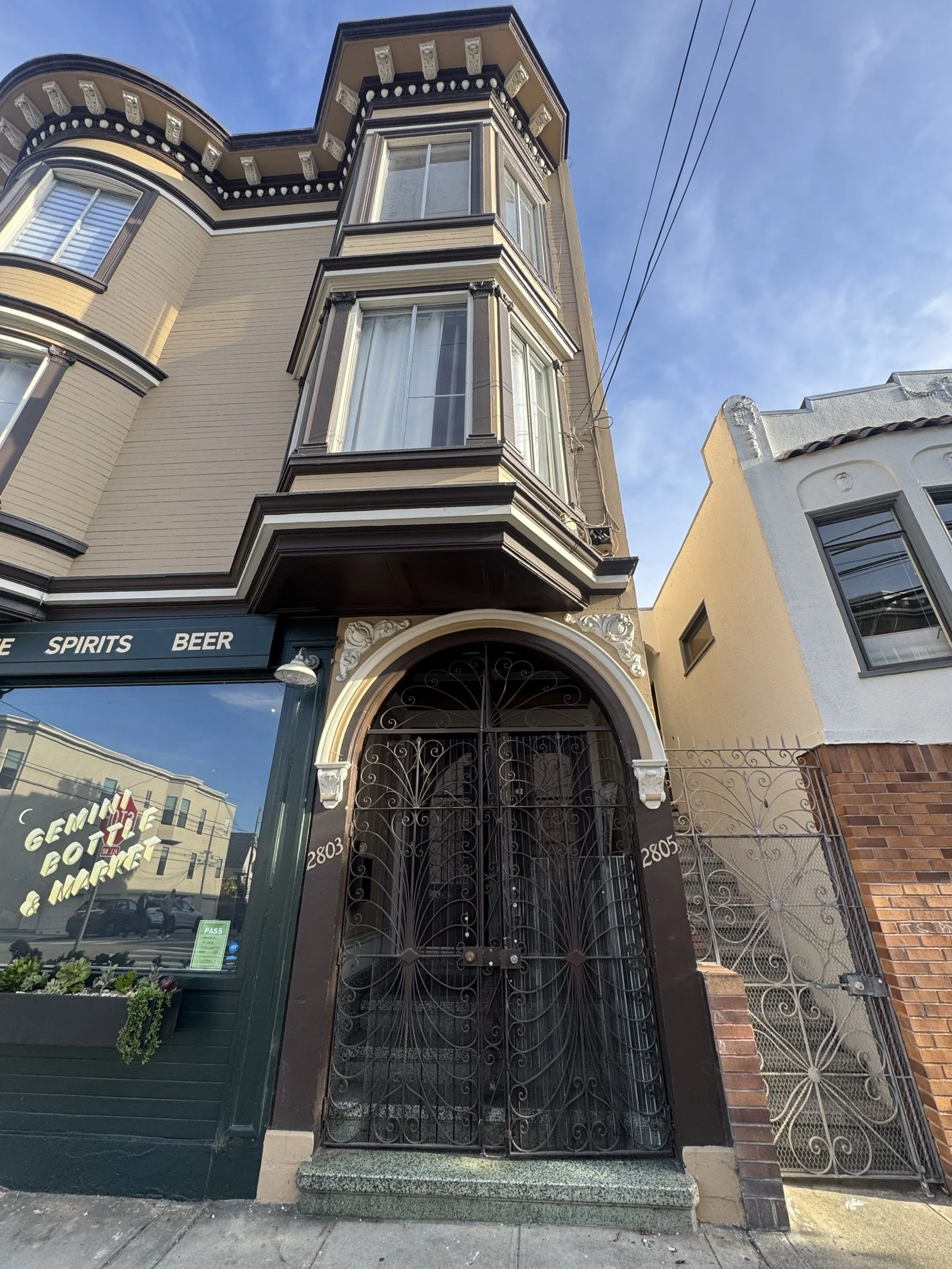 A multi-story building with decorative architectural features, including large bay windows, an ornate gate at the entrance, and a sign indicating it is a bar or pub named "Gemini, Bott & Market." The building has beige siding and black trim with whit