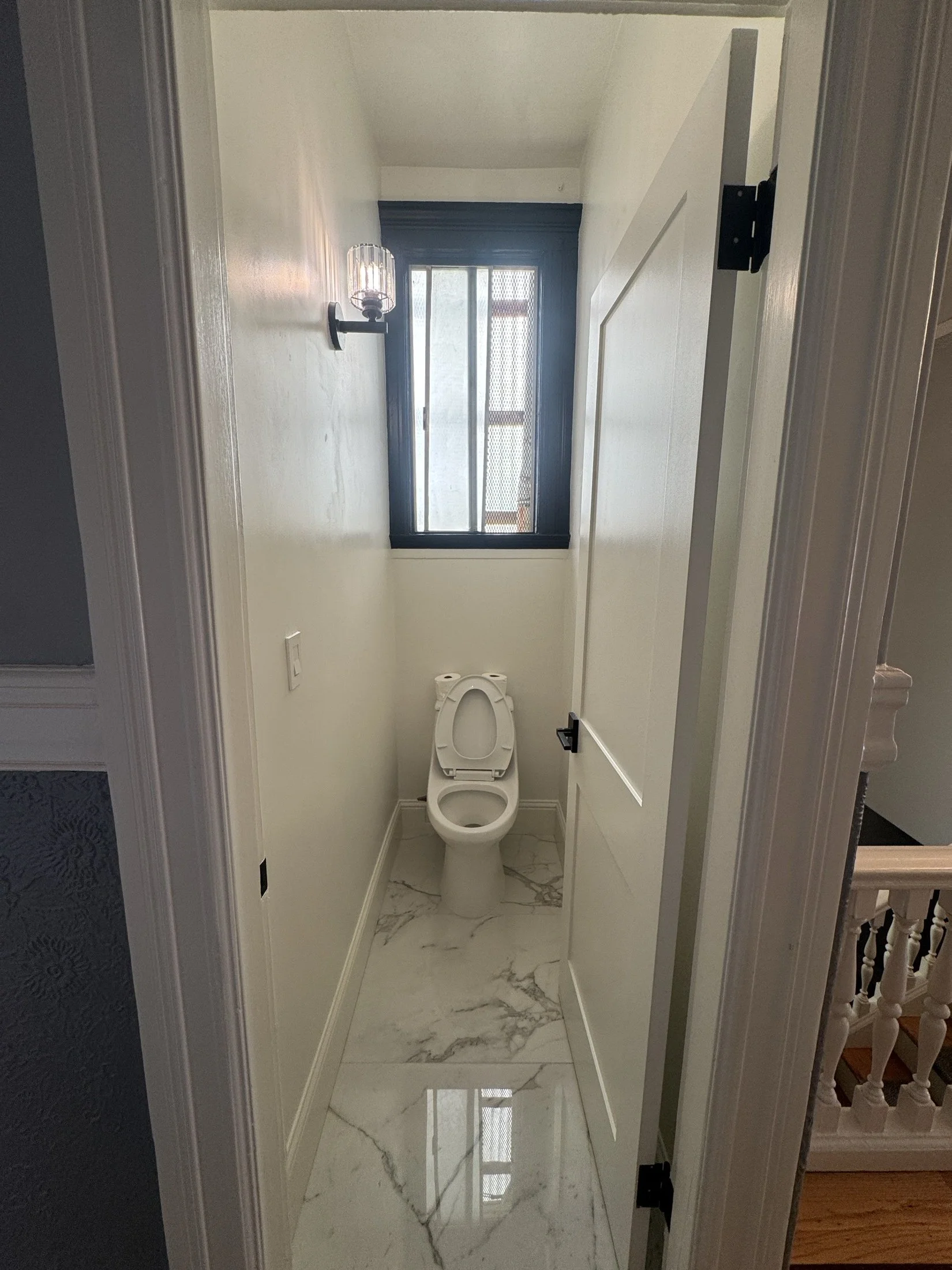 A small bathroom with a toilet, a window with screen, a wall-mounted light fixture, and marble flooring.