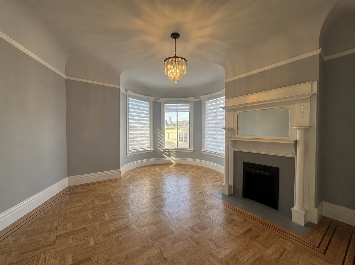Empty living room with a fireplace, hardwood floors, and a bay window with white blinds, illuminated by a chandelier.