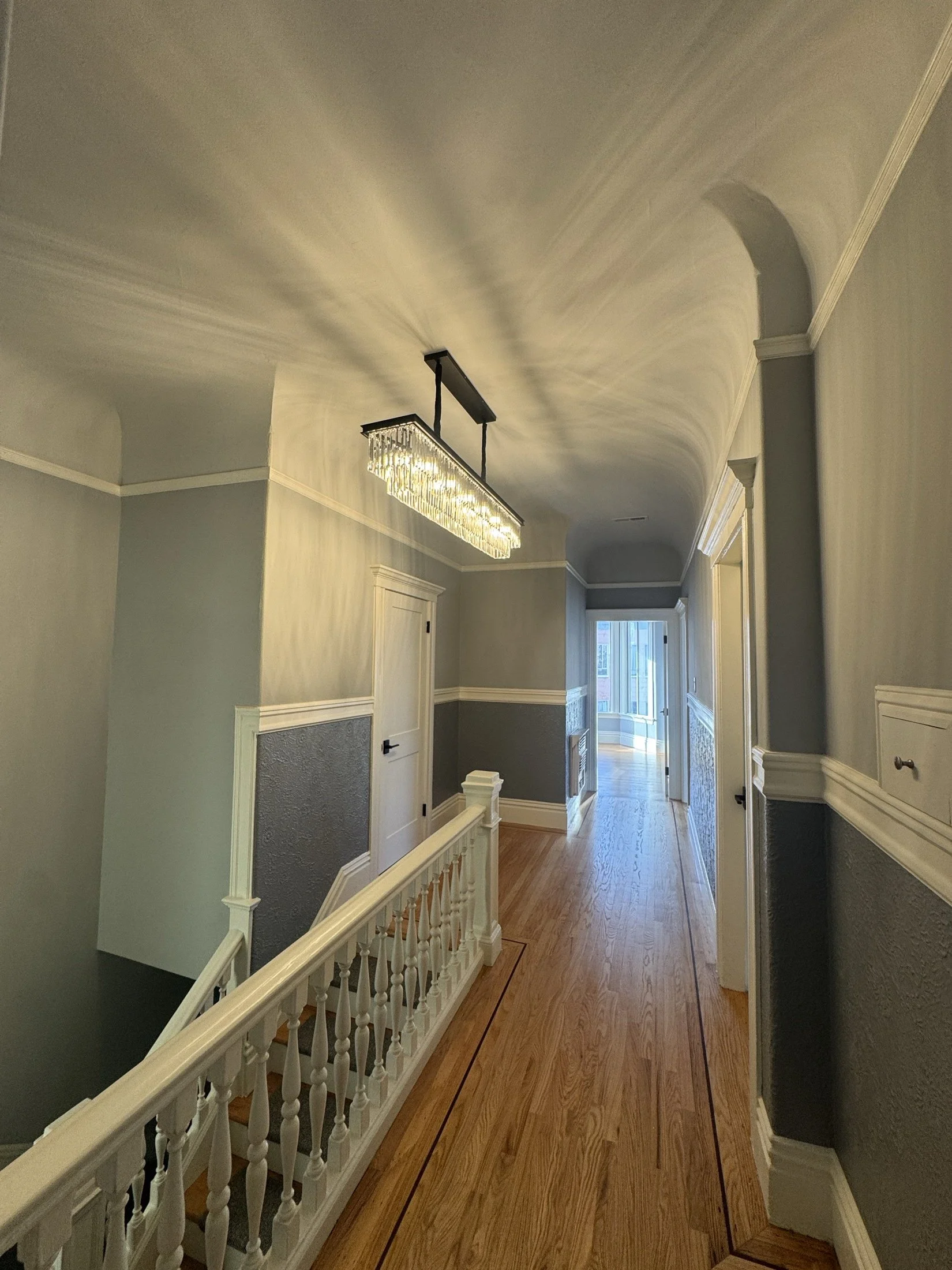 A hallway with a hanging rectangular crystal chandelier, beige and green walls with white trim, hardwood floor, windows at the end, and a white railing with spindles on the left side.