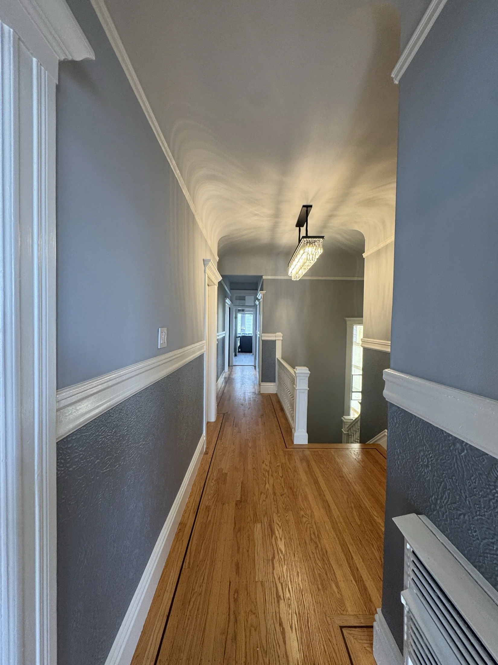 A hallway with gray walls, white trim, and hardwood floors. There are crown moldings and a hanging rectangular chandelier near the ceiling. The end of the hallway opens to a brighter room with windows.