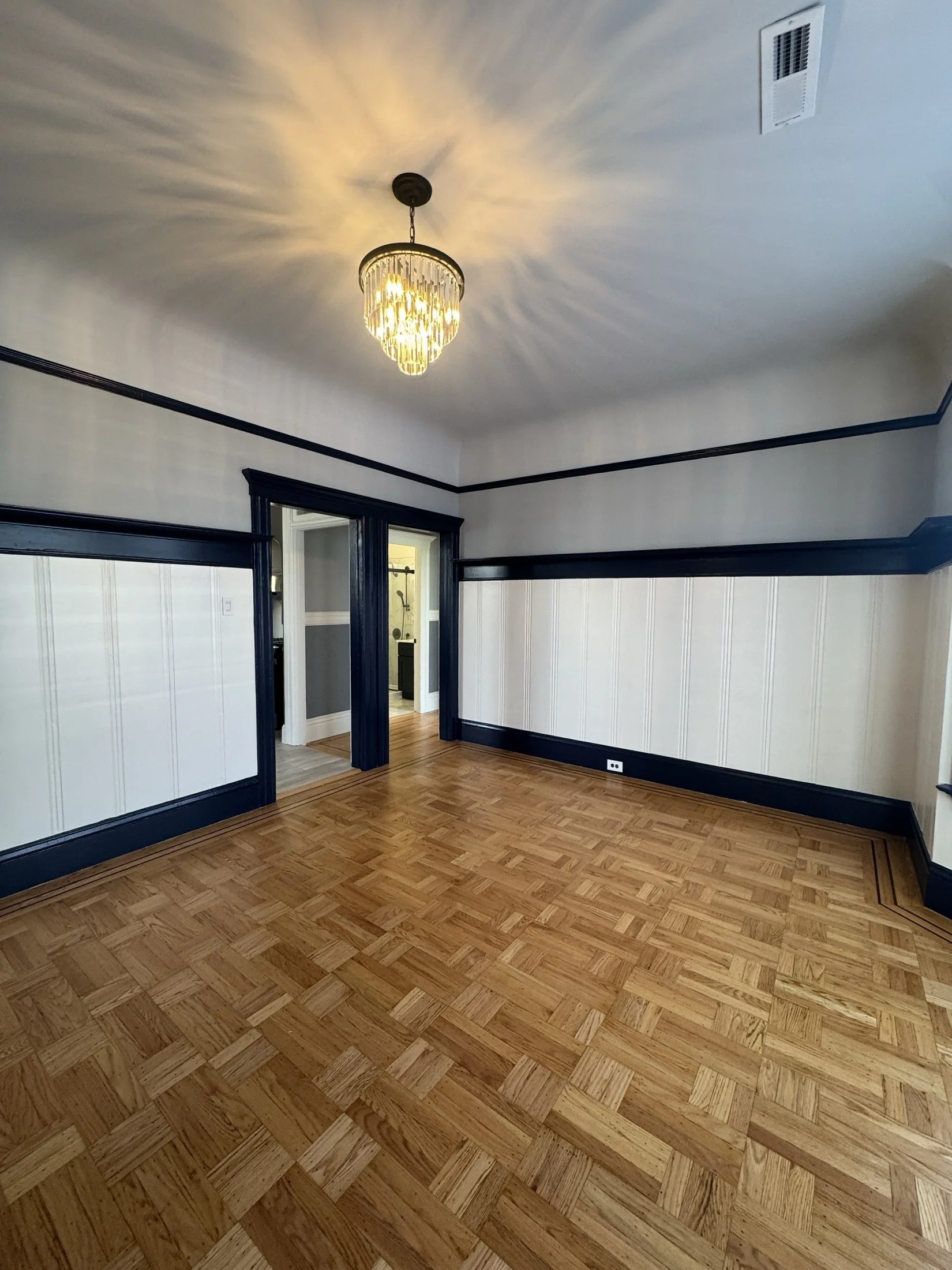 Empty room with hardwood flooring, white walls with black trim, and a chandelier ceiling light casting rays of light on the ceiling.