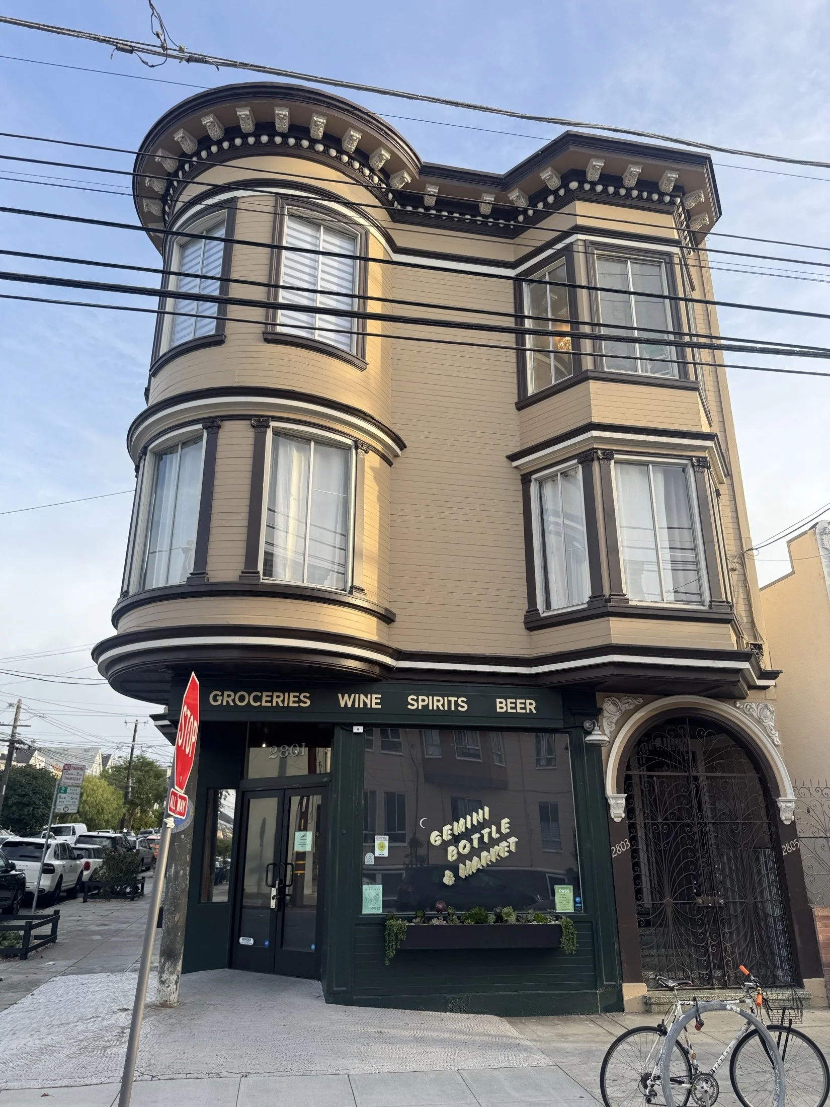 A three-story building with beige siding and black trim, featuring curved bay windows on the upper floors. The ground level has a storefront with green framing and gold lettering that reads 'Gemini Bottle & Market,' and offers groceries, wine, spirit