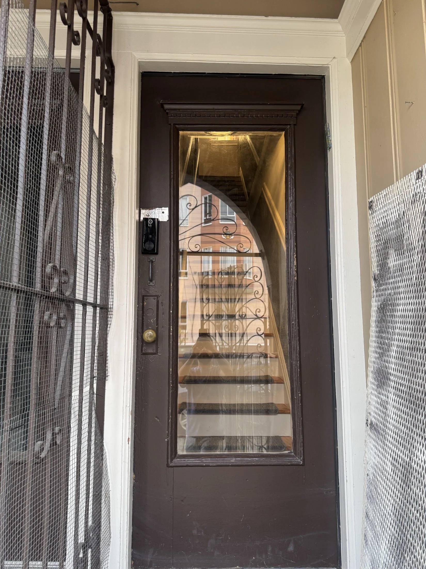 View of a building entrance with a black door, staircase reflected in the glass, insulation in a metal cage on the sides, and a small mat in front of the door.
