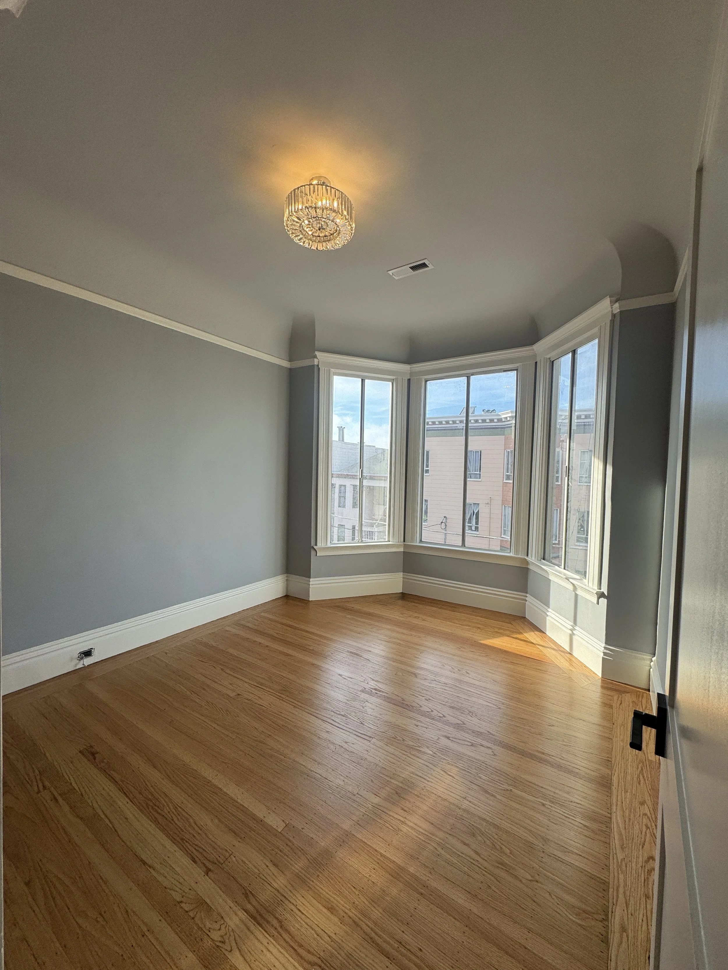 Empty room with hardwood floors, light gray walls, white crown molding, and a large bay window with a view of neighboring buildings.