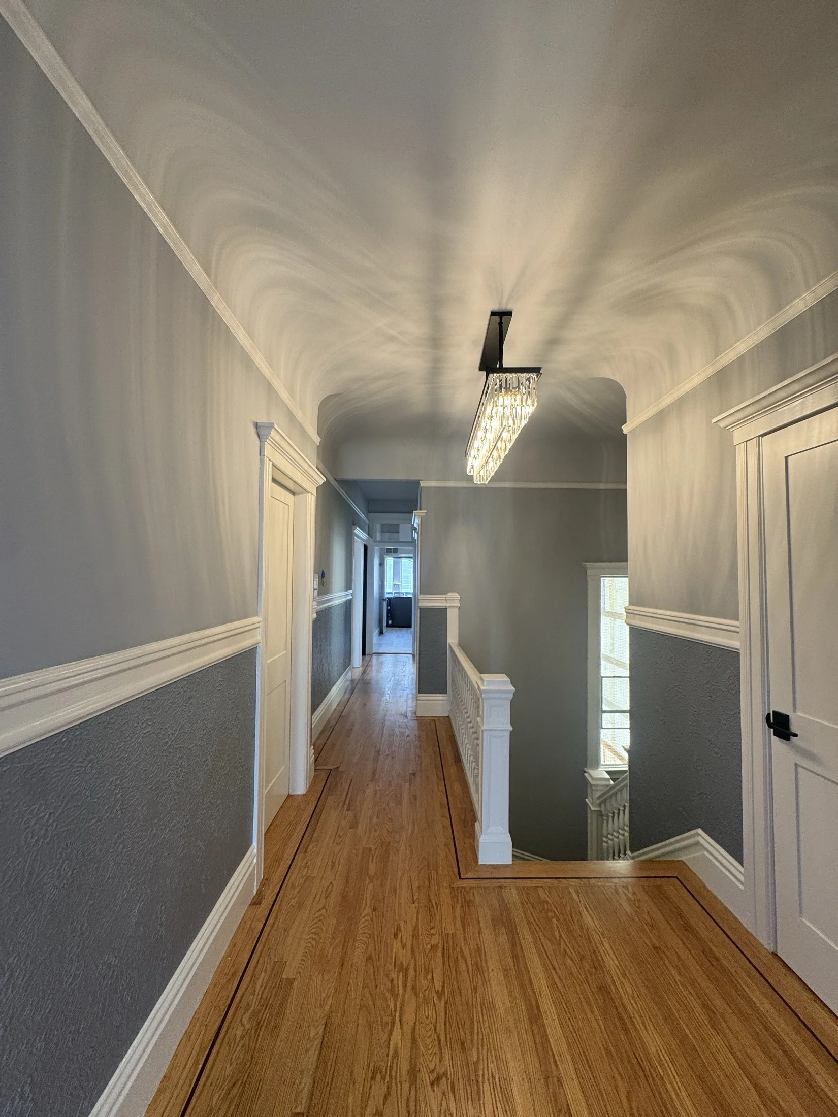 Bright hallway with wooden floors, gray wainscoting, white trim, a central rectangular modern chandelier, and staircase with white railing leading downstairs.