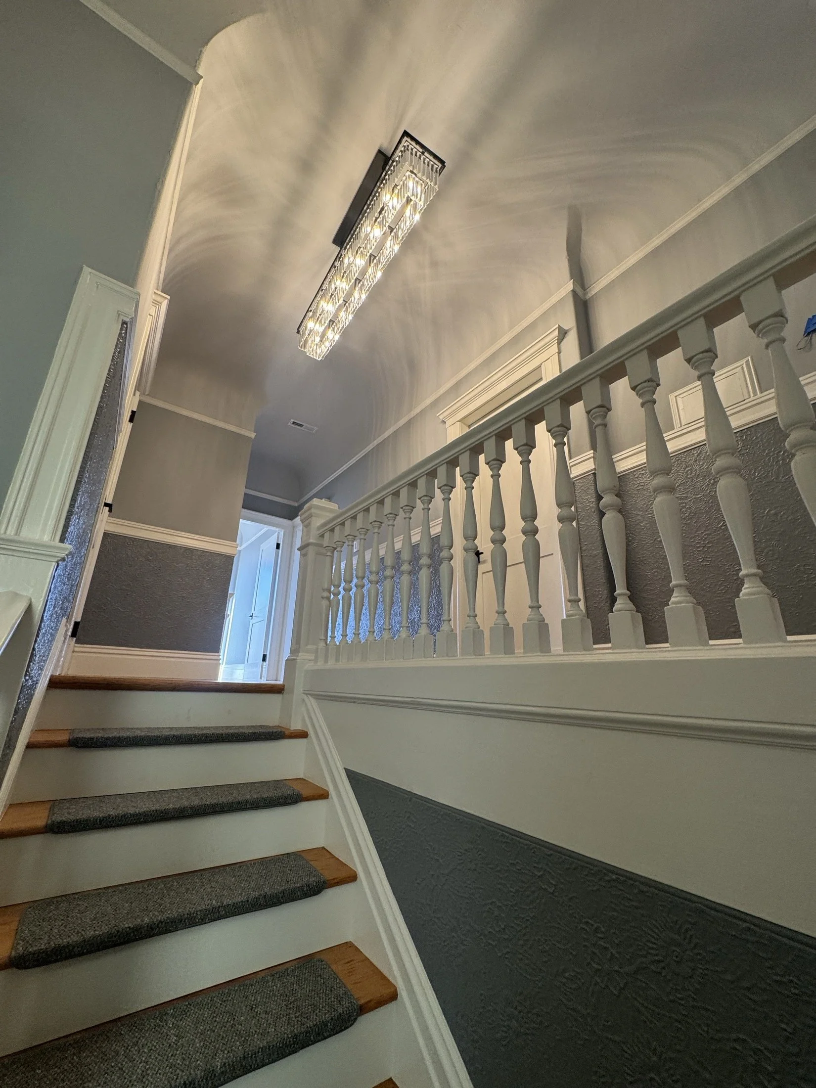 Interior view of a home staircase with gray carpeted steps, white risers, a white railing with balusters, walls painted in two shades of gray, a doorway at the top, and a decorative light fixture on the ceiling.