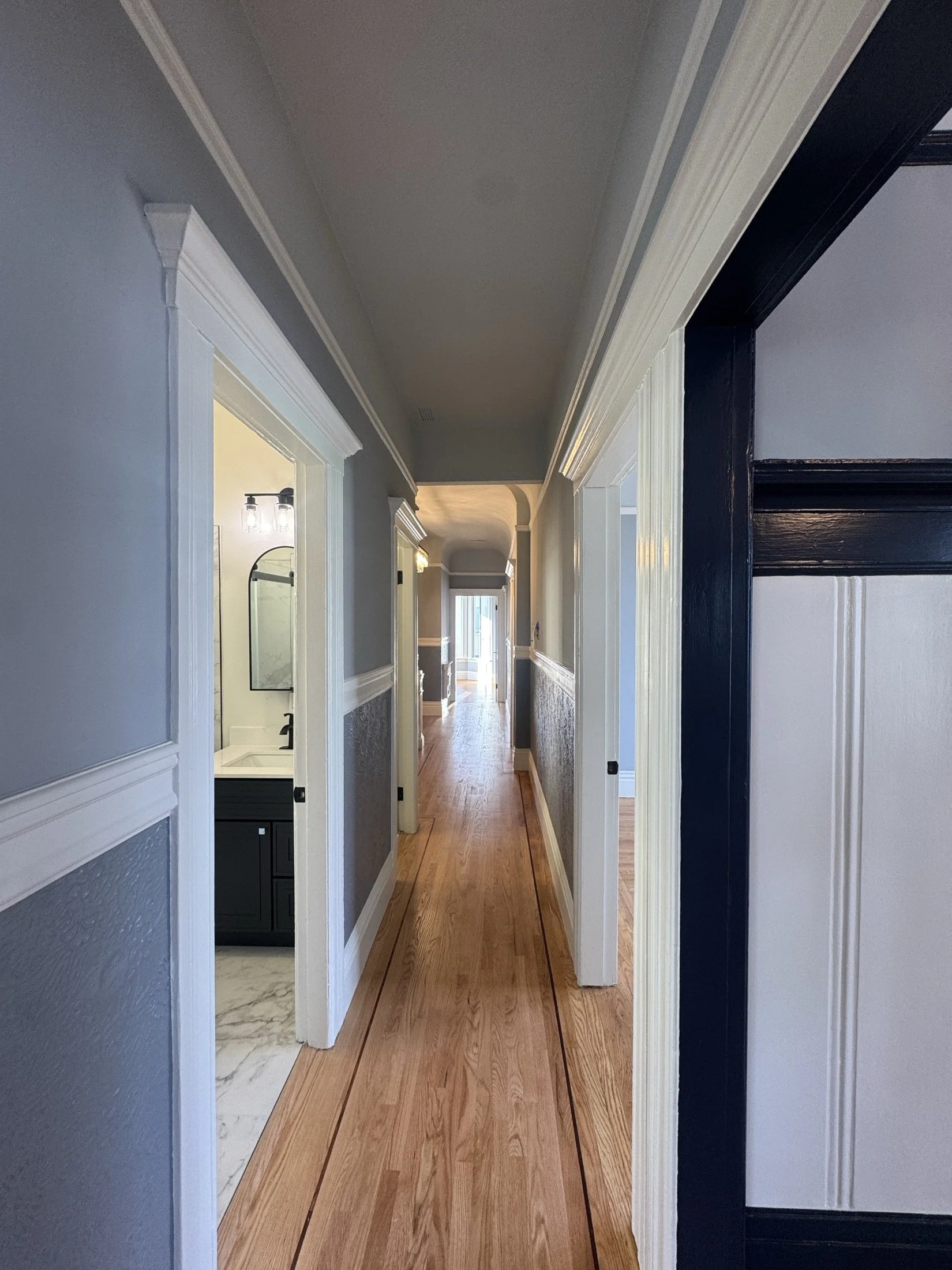A hallway inside a house with wooden flooring, white trim, and gray walls. Doors on either side lead to rooms, with a view towards a distant window at the end of the hall.