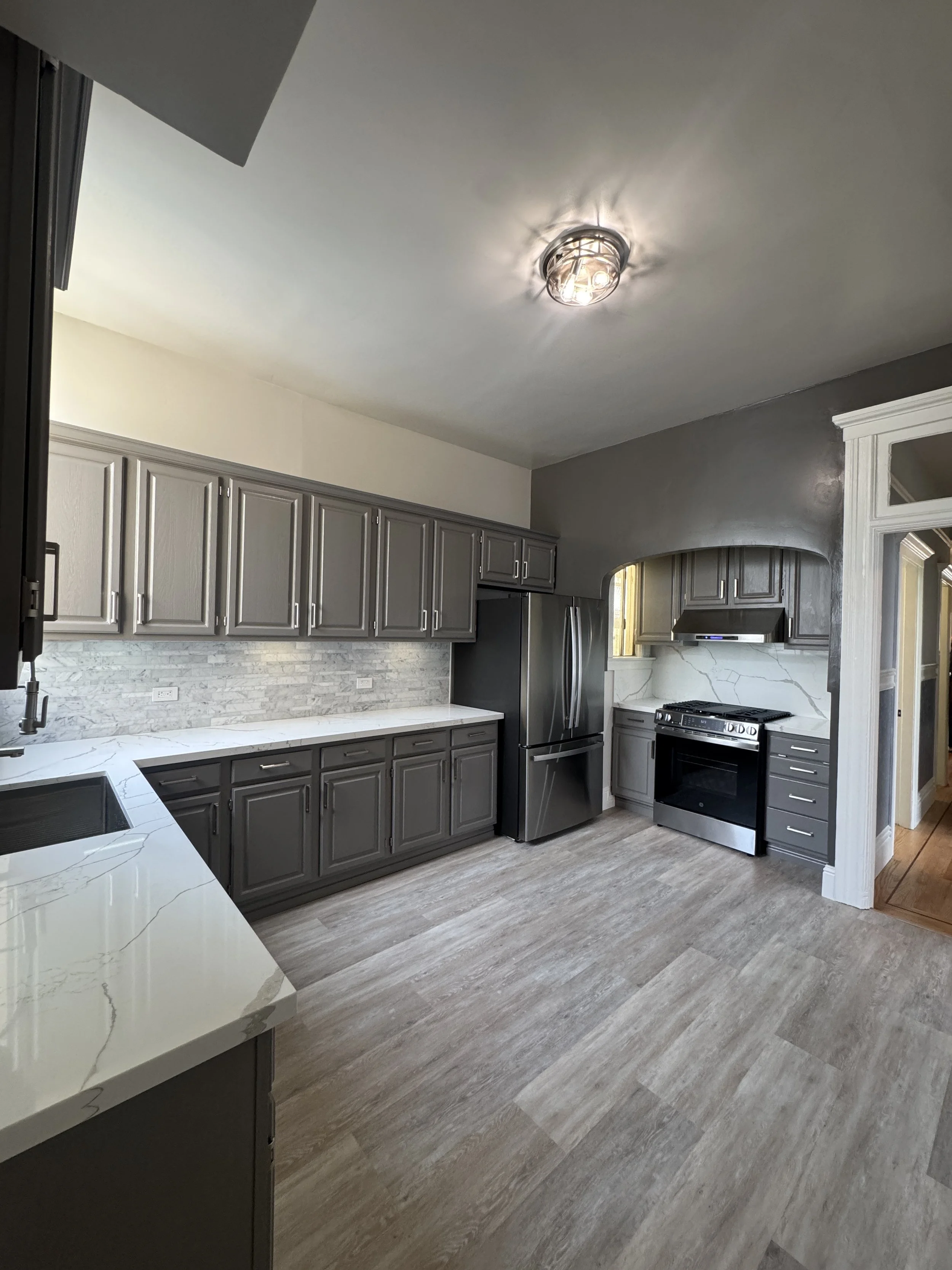 A modern kitchen with grey cabinets, stainless steel appliances, a marble countertop, and a textured tile backsplash. There is a window above the sink and a ceiling light fixture.