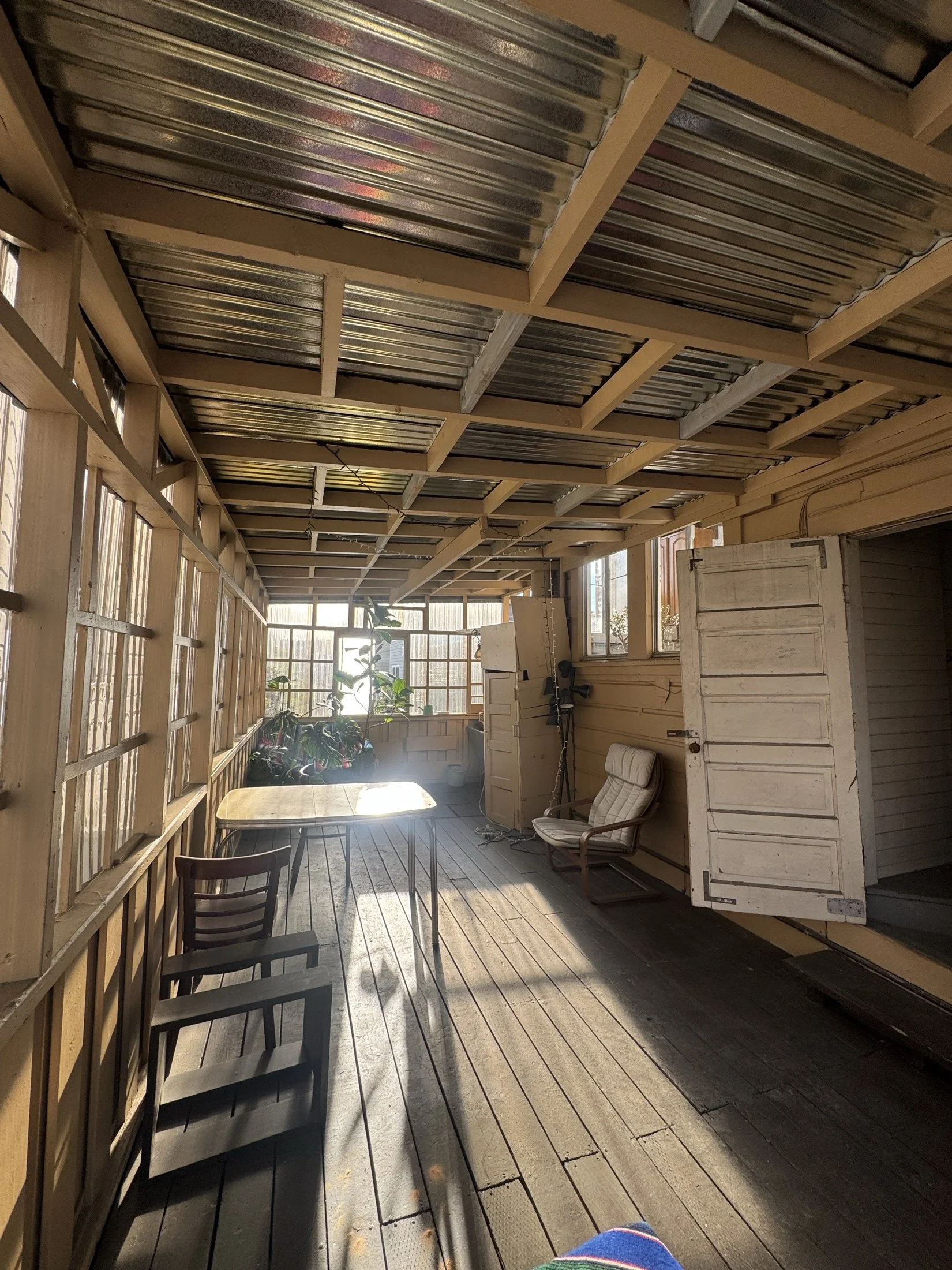 Sunlit porch or sunroom with wooden floors, chairs, a table, potted plants, and a white door, featuring windows and furniture.