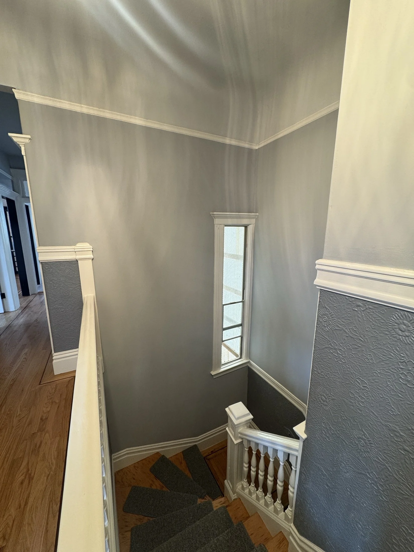 Interior view of a staircase with stained wood steps, white balustrades and newel posts, a light gray textured wall with crown molding, and a small window at the top of the stairs letting in natural light.
