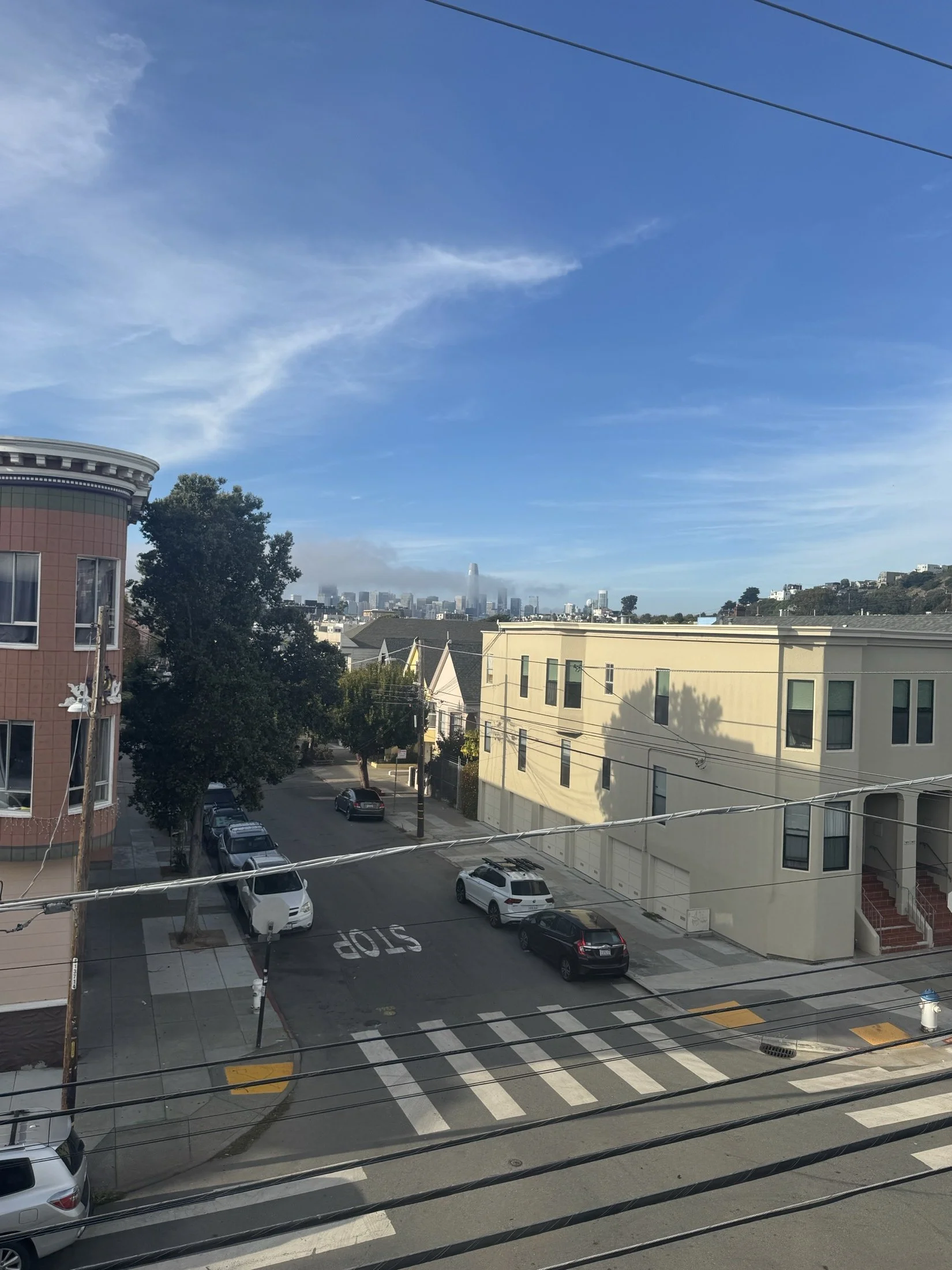 View of a cityscape with buildings, parked cars, and trees, with a clear blue sky and wispy clouds overhead.