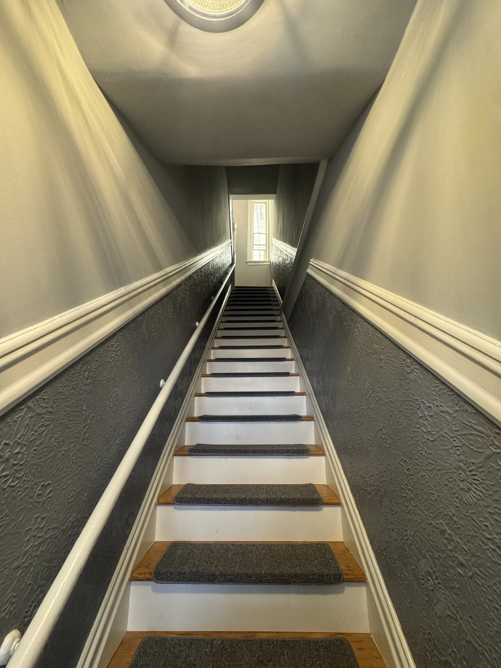 A staircase leading to a door with a window at the top, with textured dark blue lower walls, beige upper walls, a white railing, and gray steps with dark gray non-slip strips.