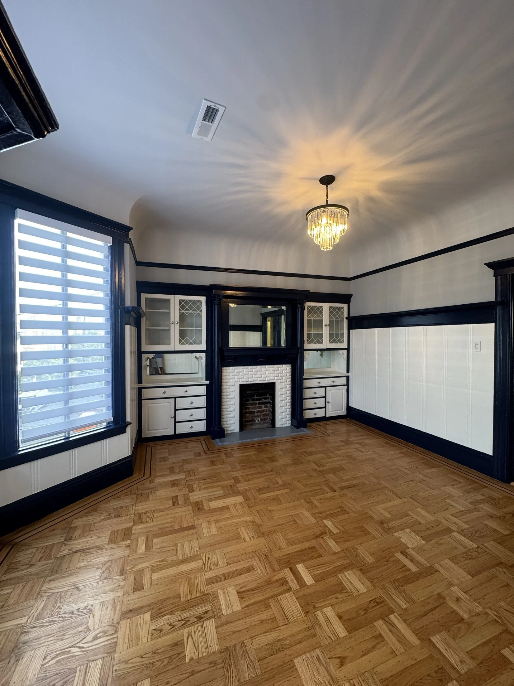 Empty living room with wooden parquet floors, built-in wall cabinets with glass doors, a fireplace with a brick surround, a window with horizontal blinds, a chandelier on the ceiling, and dark picture frame moldings.