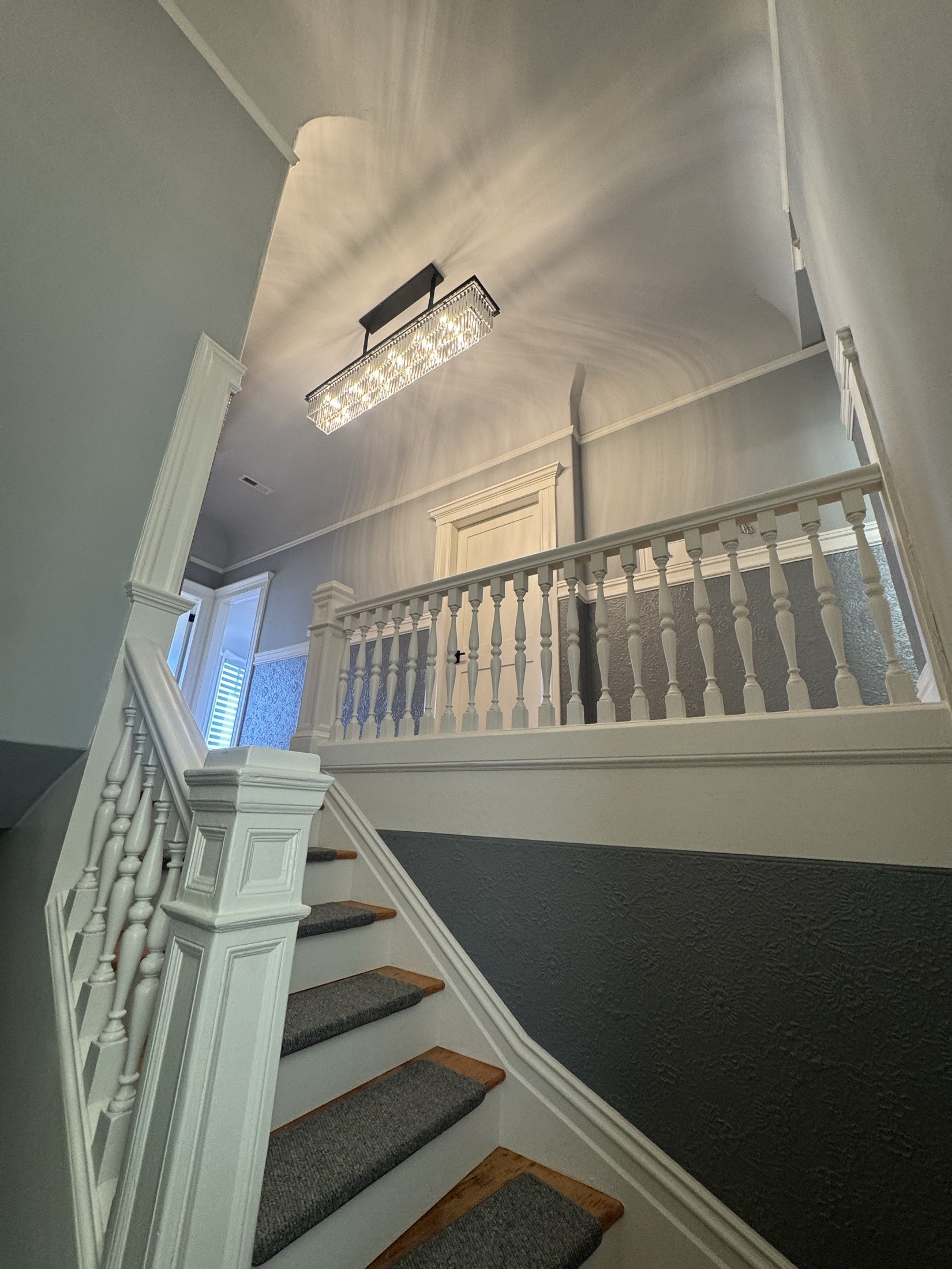 Interior view of a staircase with gray carpeted stairs, white wooden balusters, a dark textured wall, and a hanging rectangular crystal chandelier casting light and shadow patterns on the ceiling.