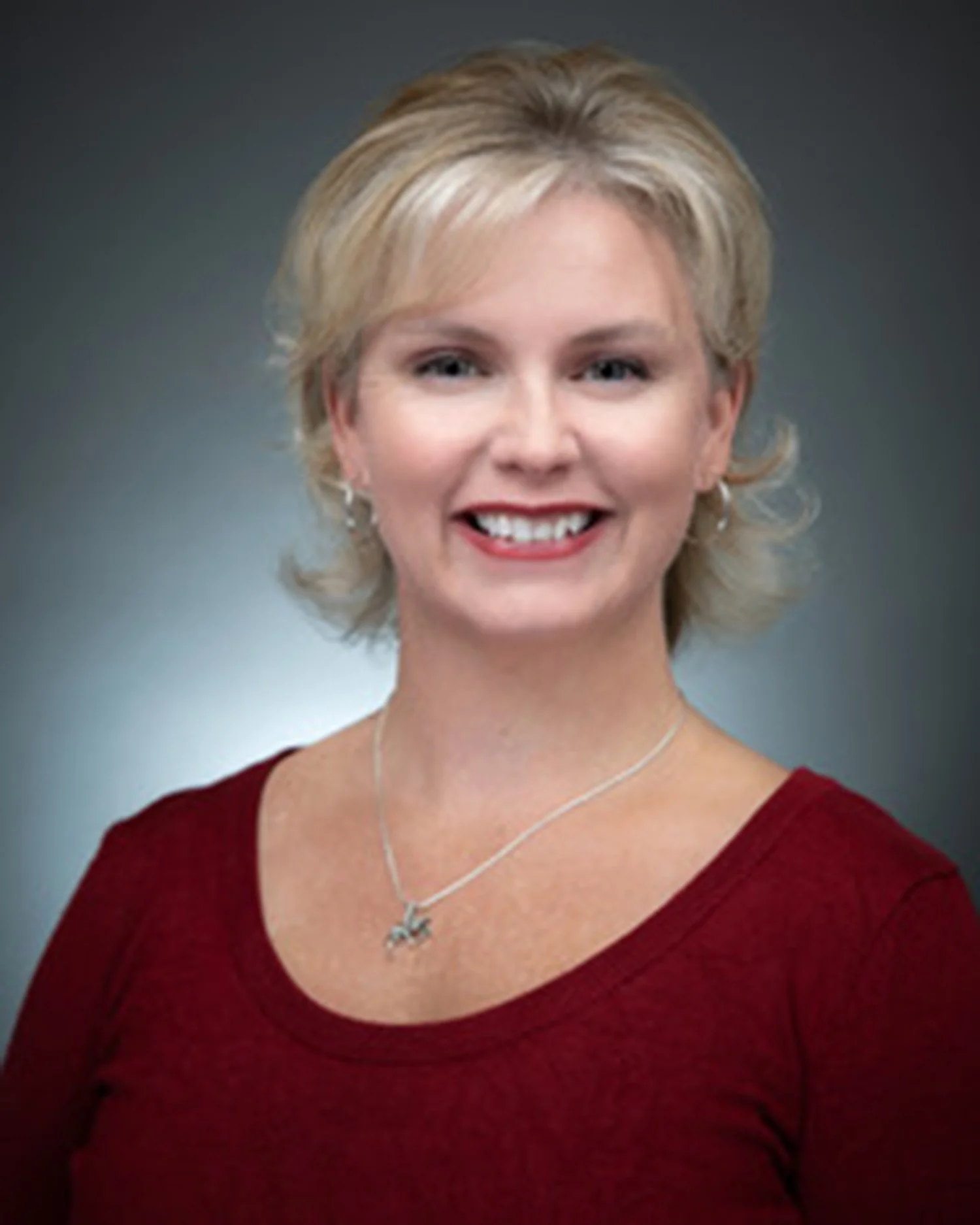 Headshot of a woman with short blonde hair, wearing a red top, silver necklace, and earrings, smiling against a gray background.