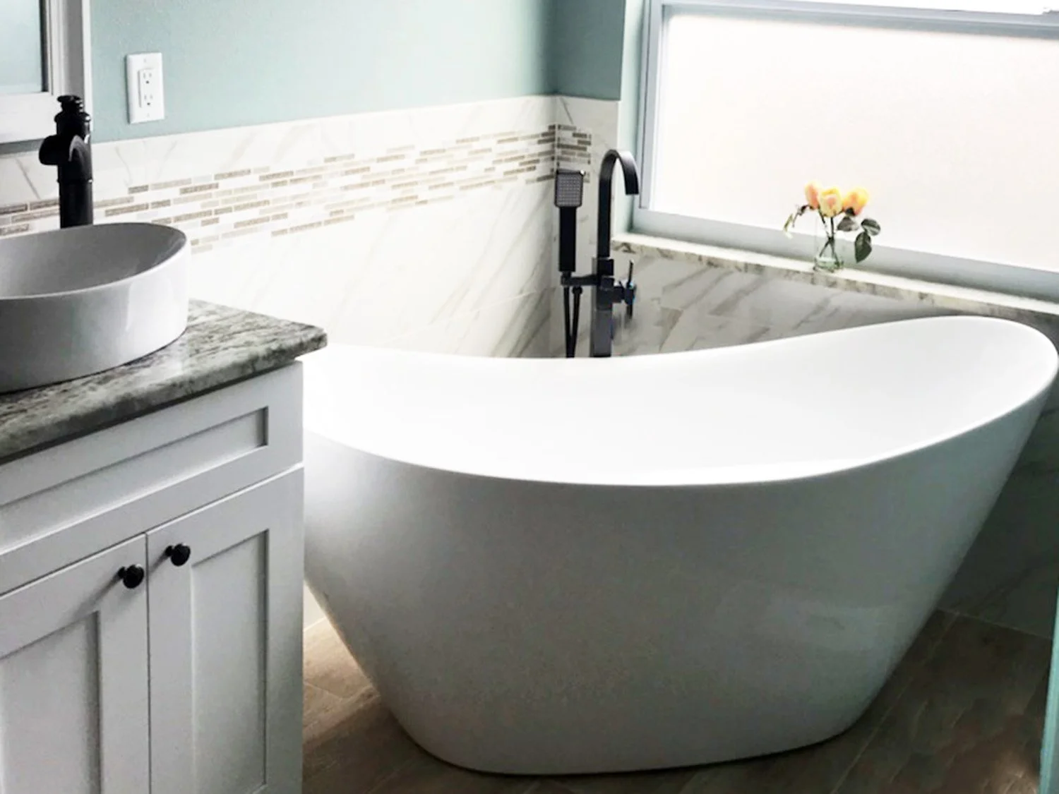 Bathroom with a freestanding bathtub near a window with a small vase of yellow flowers on the windowsill, a marble bathroom counter with a vessel sink, and a black faucet.