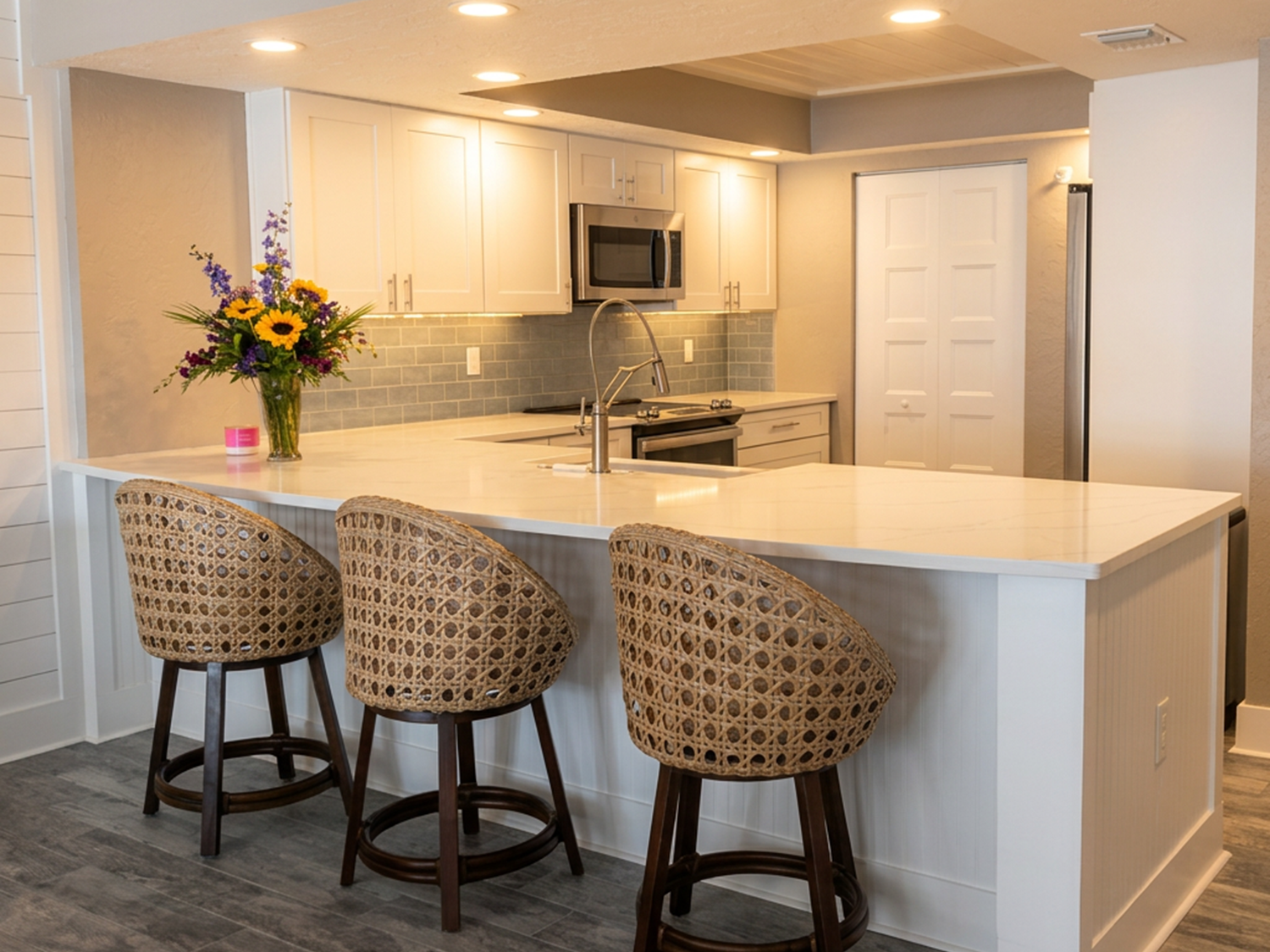 Kitchen with white cabinets, gray backsplash, stainless steel microwave, stove, and refrigerator. A large island with a white countertop has a vase of colorful flowers and three woven barstools.